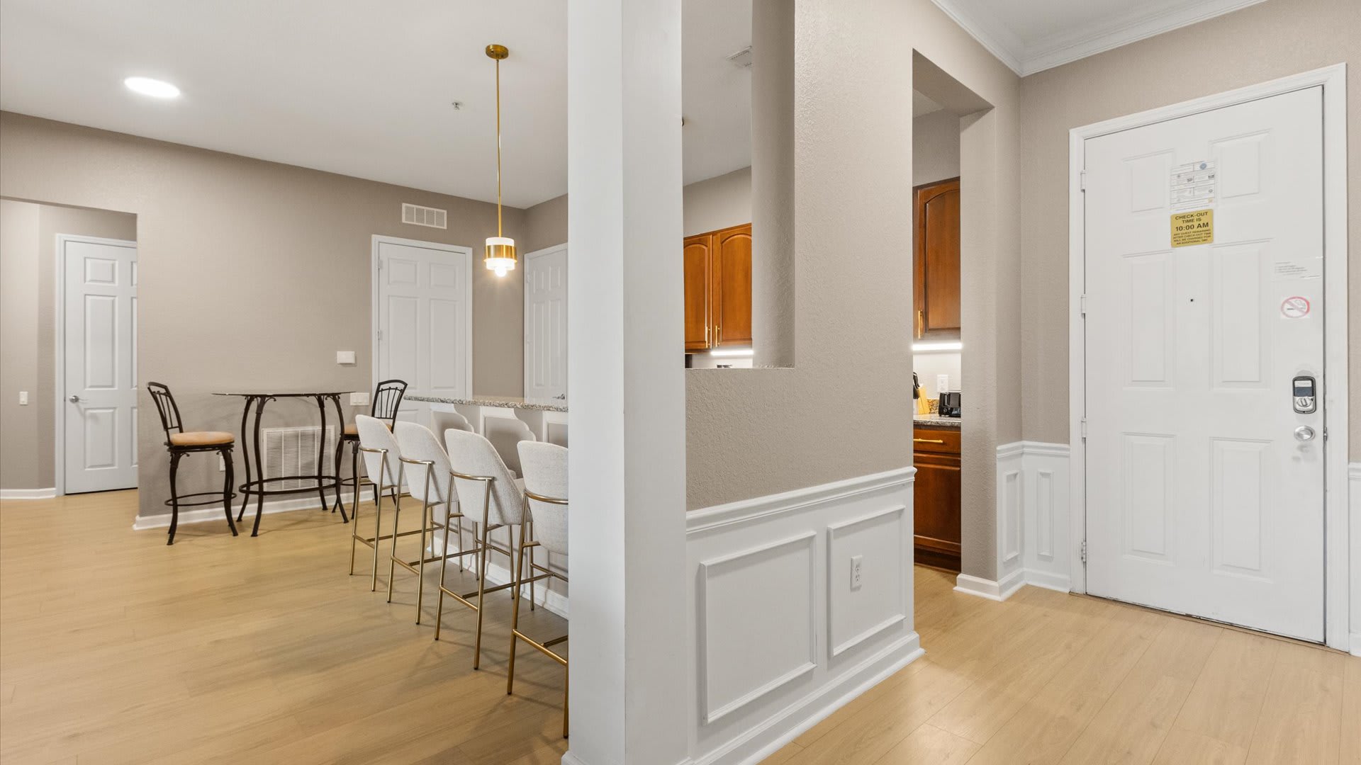 A view from the entryway showing a partial kitchen island with bar stools and a small round bistro table with chairs in the background. Light wood floors extend throughout the open-concept space, with a white door on the right and warm-toned kitchen cabinets visible.