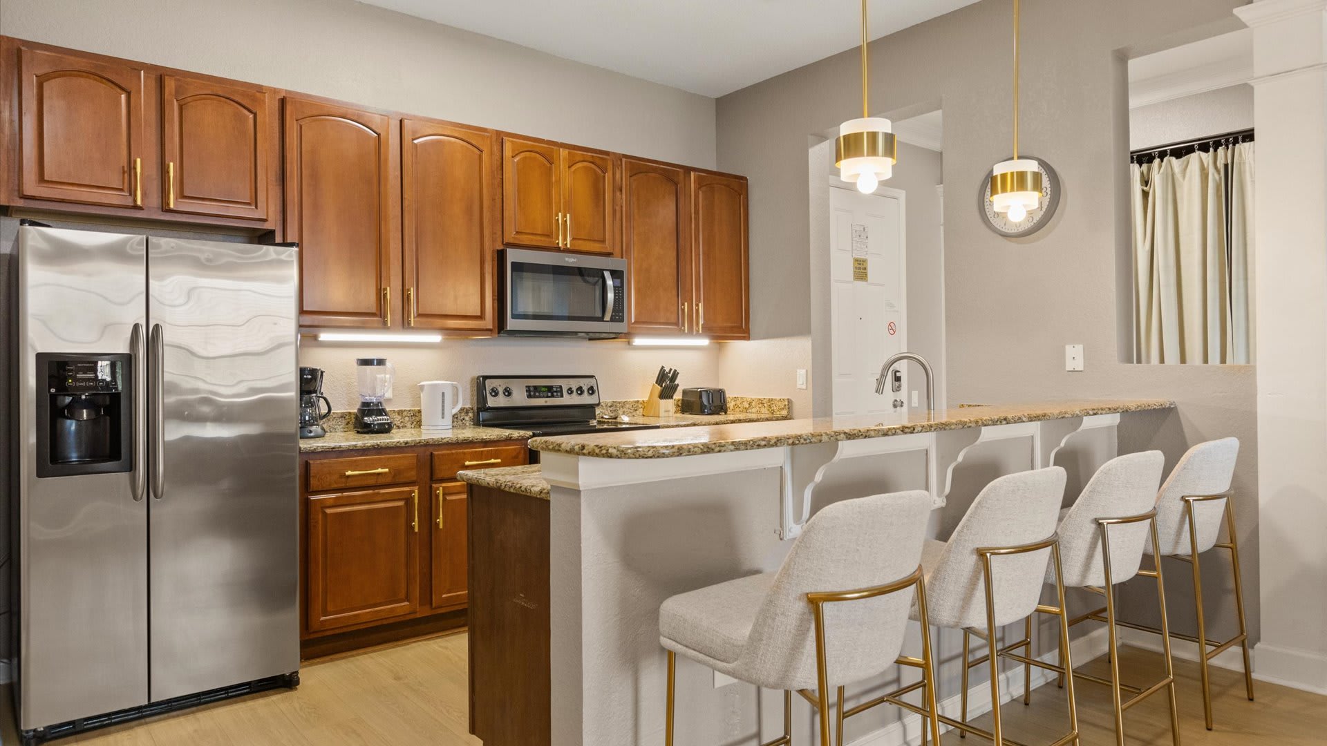 A wide shot of the kitchen showcasing the breakfast bar with four bar stools, granite countertops, and warm wood cabinetry. Stainless steel appliances, including a refrigerator and microwave, are visible, along with two modern pendant lights above the bar.