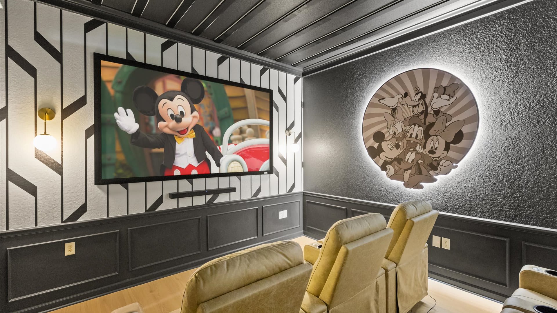 Dark-themed media room featuring a large projection screen displaying Mickey Mouse, two tan leather recliners, and a circular backlit Disney character art piece on the textured wall.
