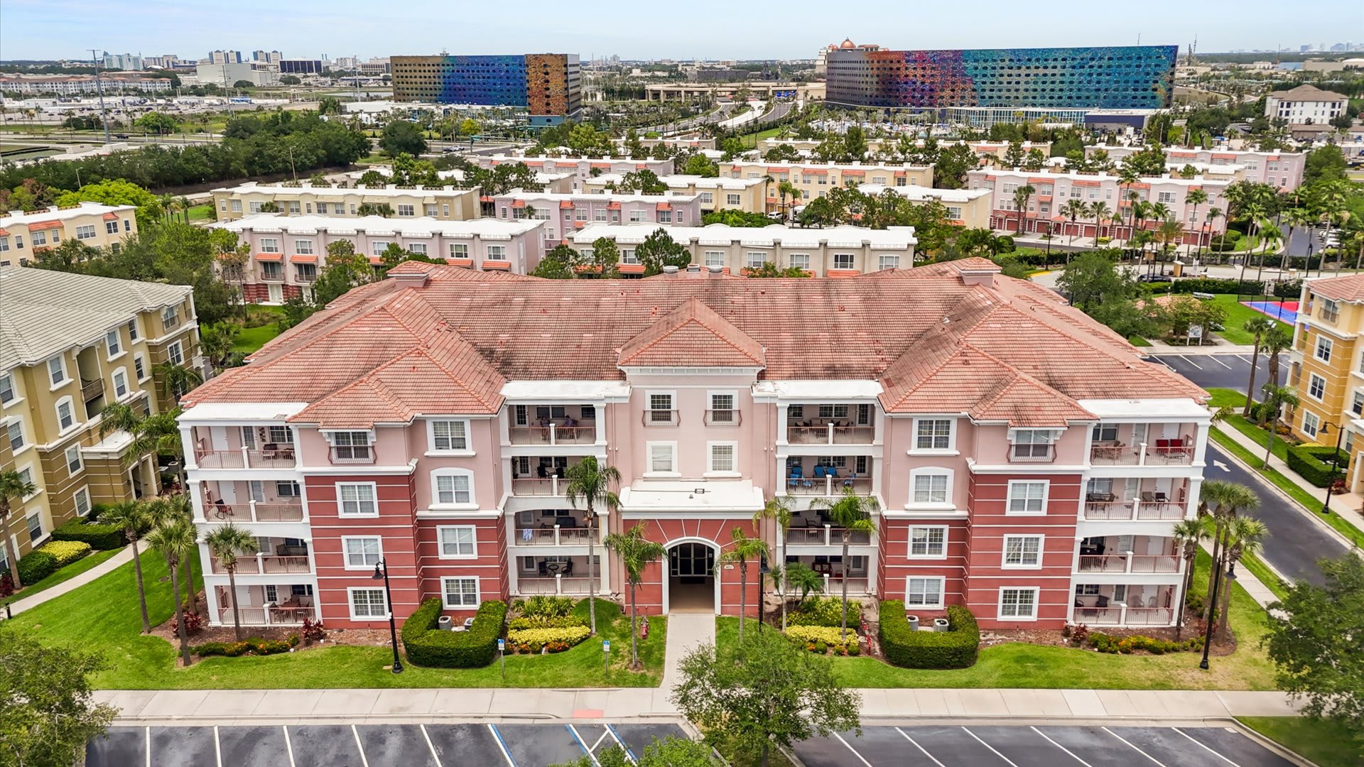 Elevated aerial view showcasing the full facade of a large, multi-story condominium building with a red tile roof and pink/red exterior, nestled among other residential complexes and distant city structures.