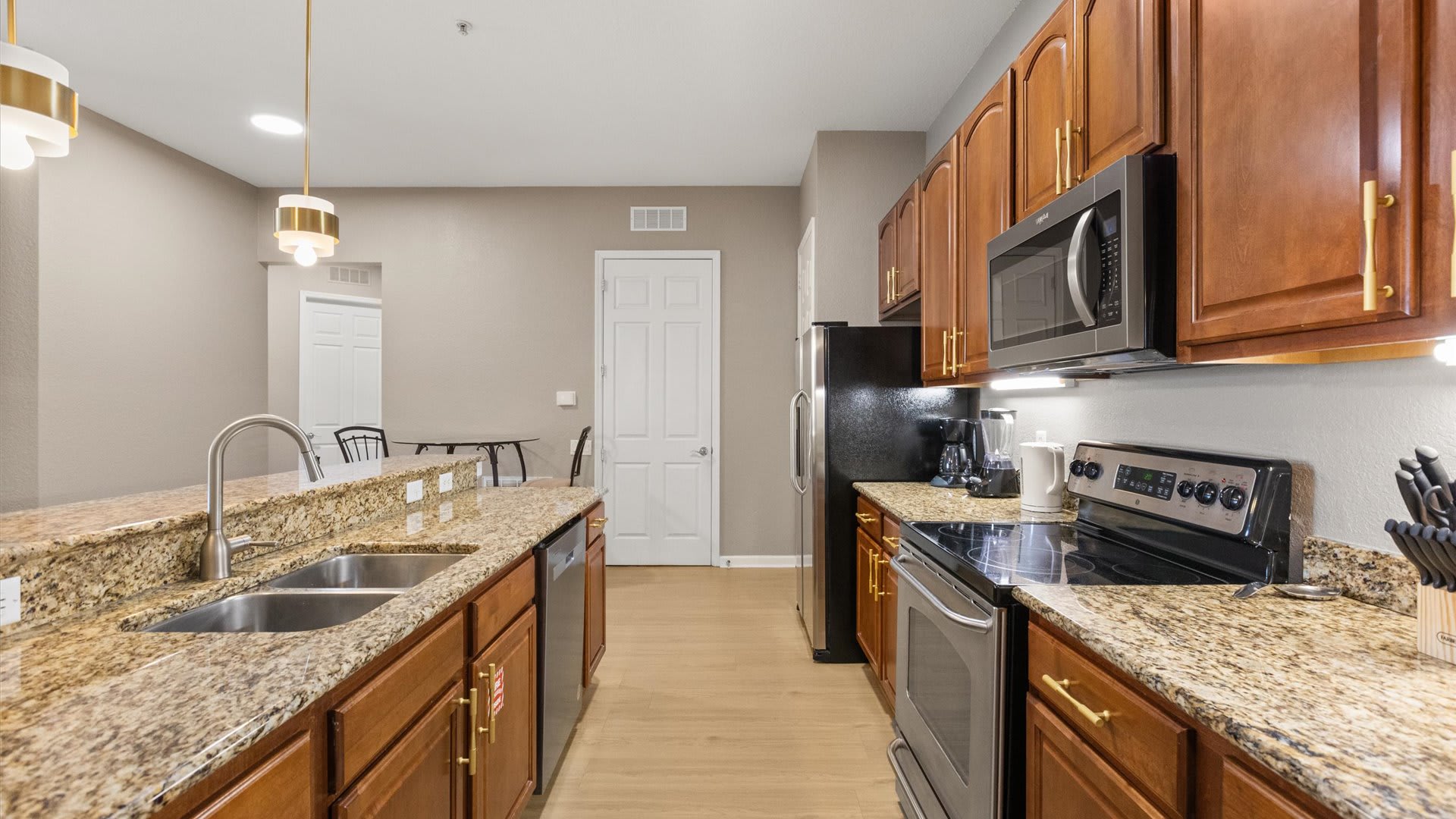 A galley-style kitchen with warm wood cabinets, granite countertops, and black and stainless steel appliances. The view shows the sink area on the left and the stove/oven on the right, with light wood floors and a white door in the background.