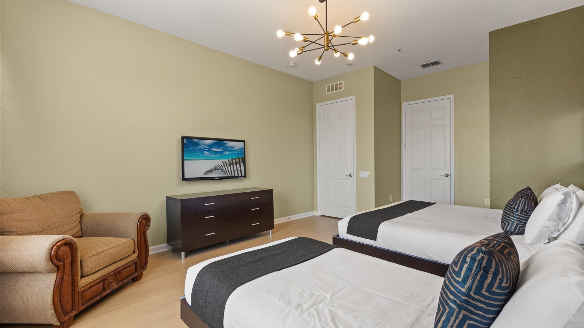 A spacious bedroom with light beige walls, two twin beds with dark grey runners, a dark wood dresser with a TV, and a modern sputnik chandelier. A brown armchair sits in the corner.