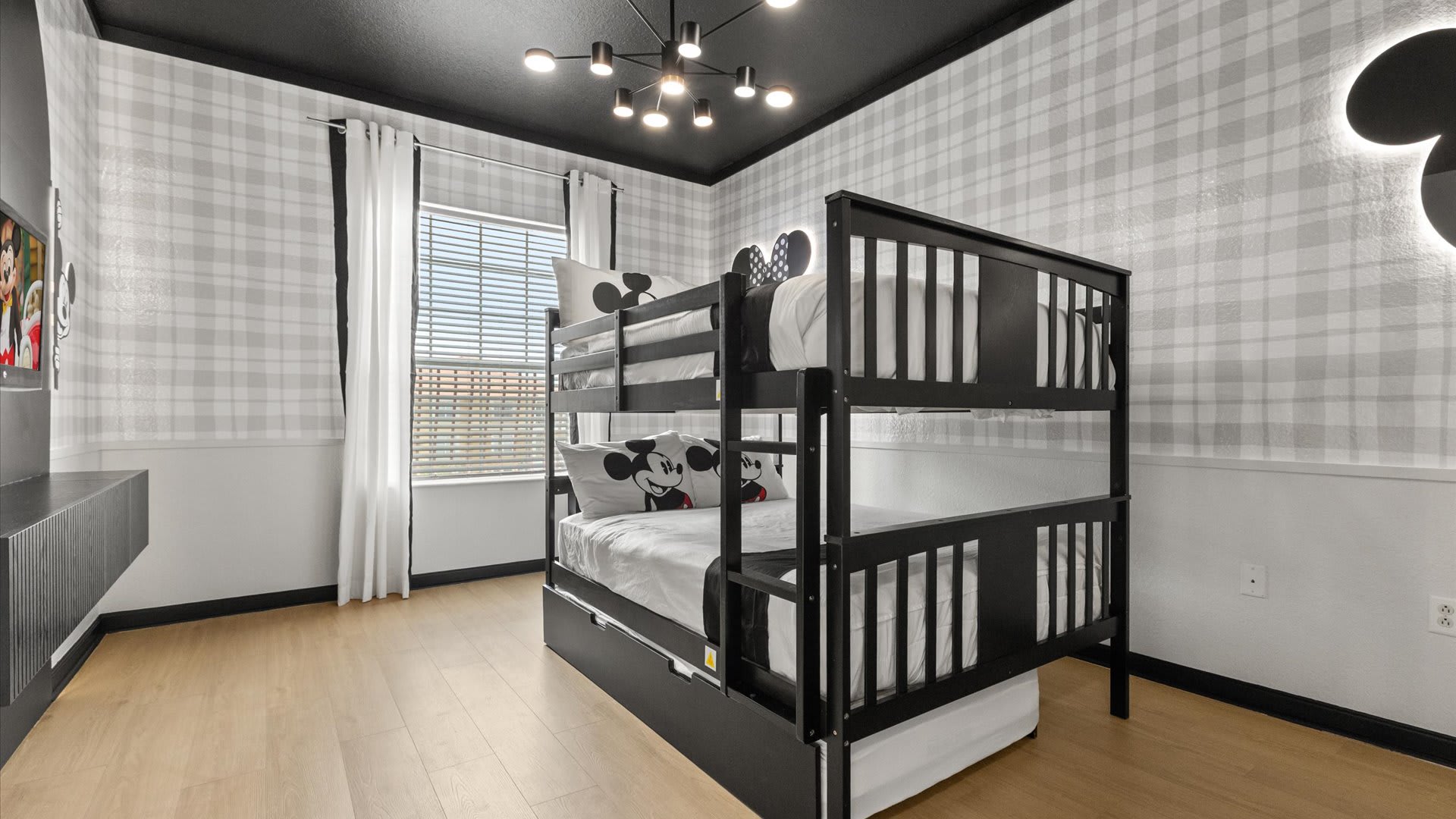 A themed children's bedroom featuring a black bunk bed with Mickey Mouse themed pillows, plaid wallpaper, and a dark ceiling with a modern chandelier. A window with blinds is visible.