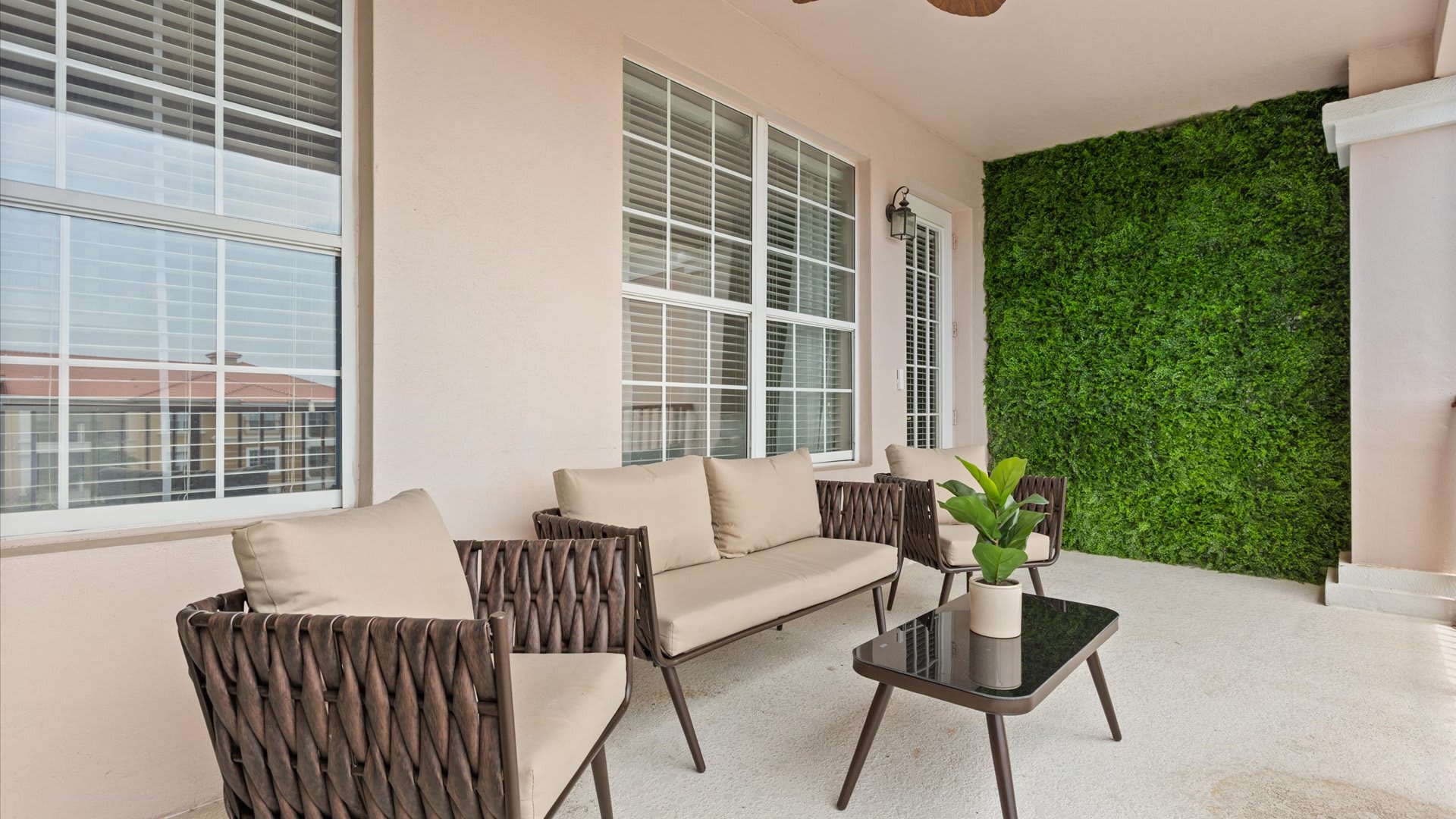 Covered balcony with a green artificial turf wall, outdoor sofa, armchairs, and a coffee table. Large windows and a door lead back into the property.