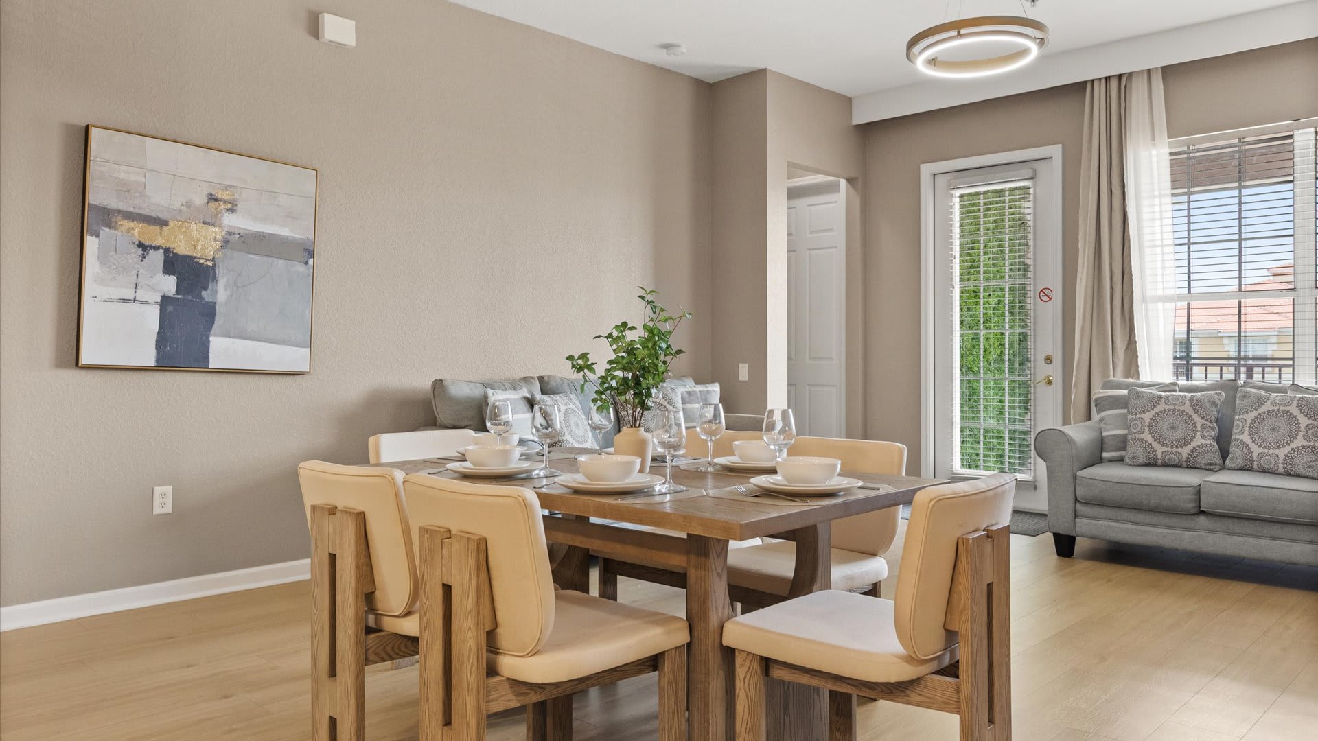 A modern dining area features a light wood rectangular table set for six with white dishes and wine glasses, complemented by light beige upholstered chairs. An abstract painting hangs on the neutral wall, and a living room with a gray sofa is visible through a doorway.