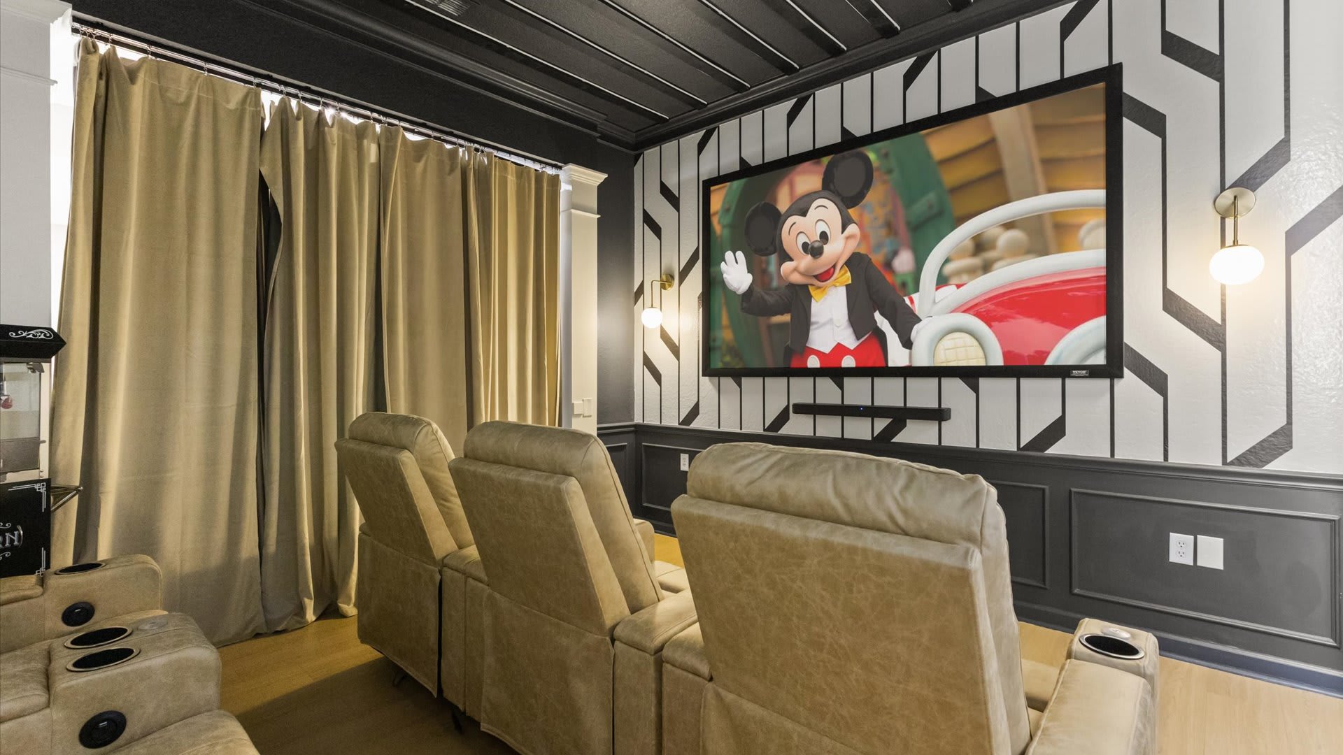Media room with a large projection screen, multiple reclining theater seats, and heavy gold curtains on one side. The ceiling features black and white striped panels with integrated lighting.
