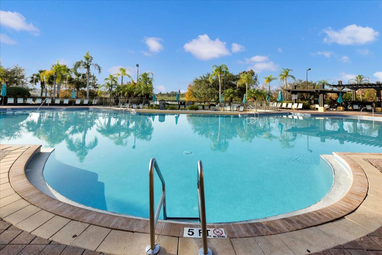 Expansive outdoor swimming pool with a curved edge, surrounded by a brick-paved deck, lounge chairs, and palm trees under a bright blue sky with scattered clouds.