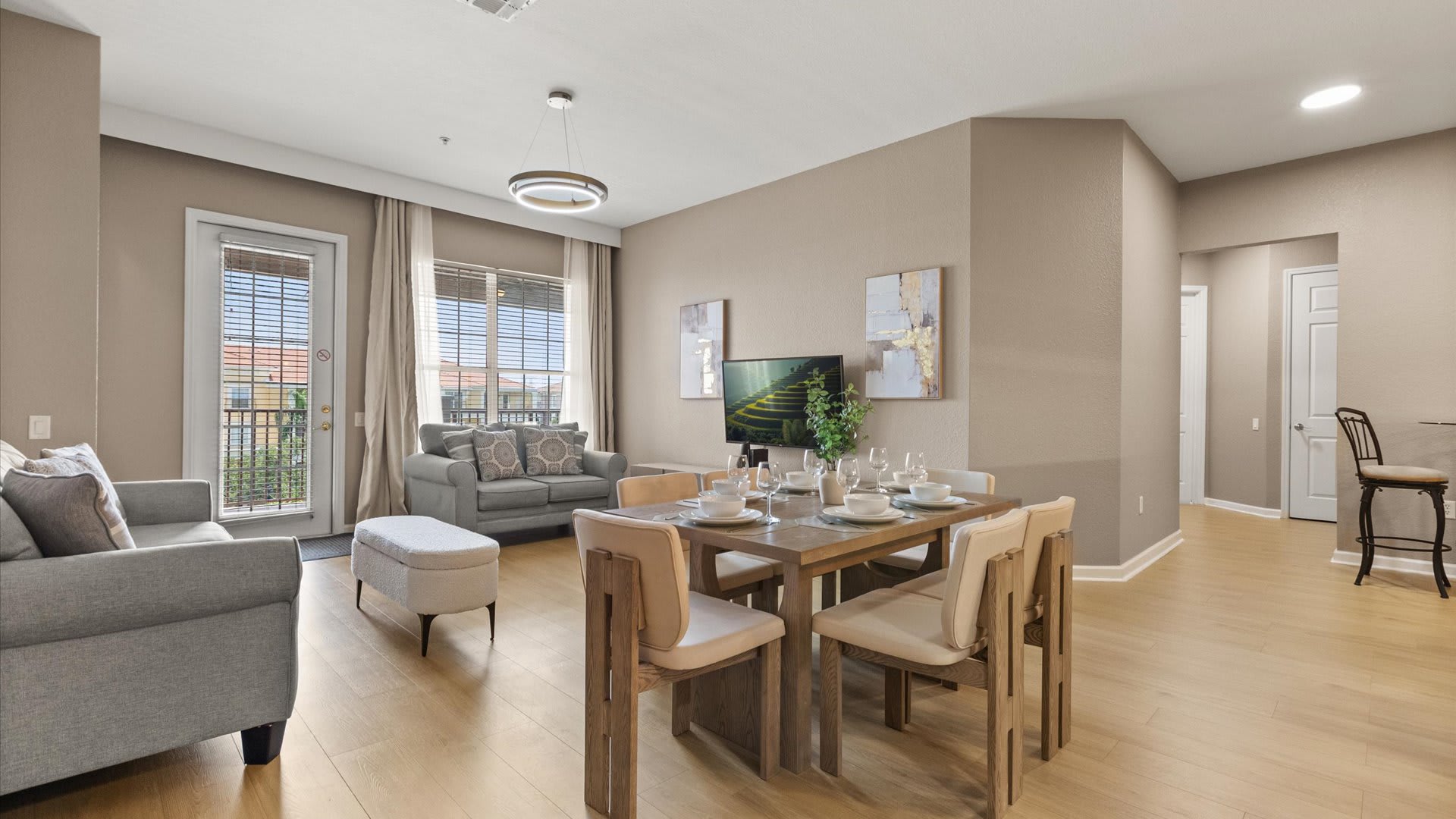 Bright open concept living and dining area with light wood floors, a gray sofa, a dining table with light wood chairs, and access to a balcony through a glass door.