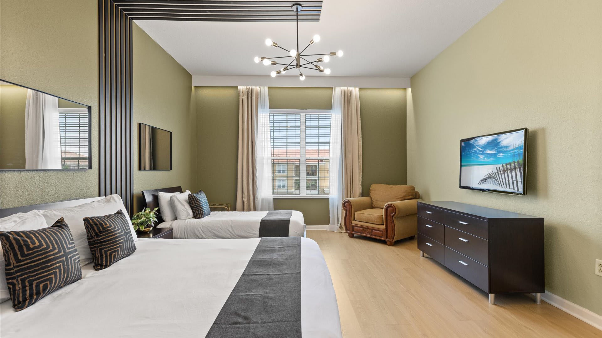 A spacious bedroom with light green walls and light wood floors, featuring a large bed and a twin bed, both with white and gray bedding. A modern sputnik chandelier hangs from the ceiling, and a flat-screen TV is mounted on a dark dresser opposite the beds.