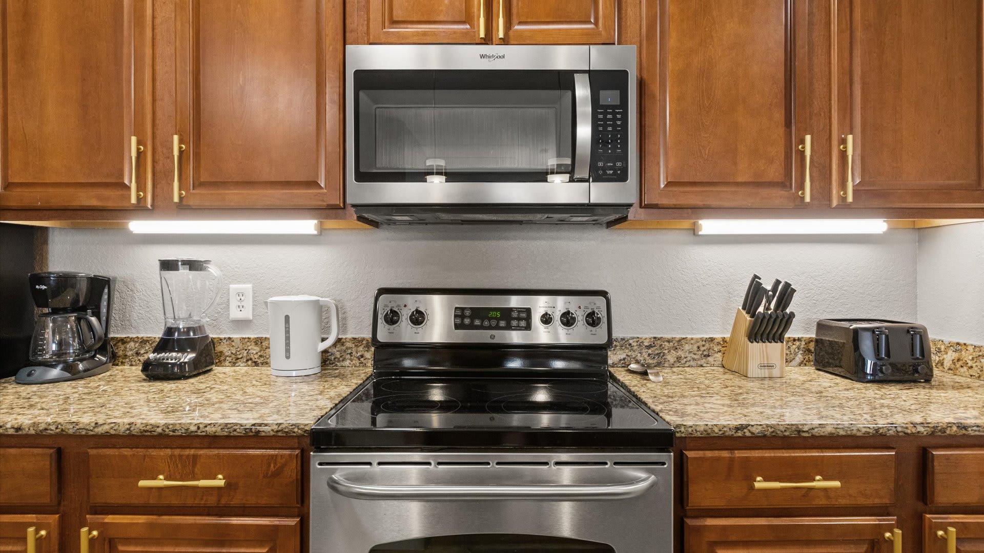 A close-up of the kitchen counter area, featuring a stainless steel range with a microwave above it, and various small appliances like a coffee maker, blender, kettle, and toaster. The granite countertops and warm wood cabinets are well-lit by under-cabinet lighting.