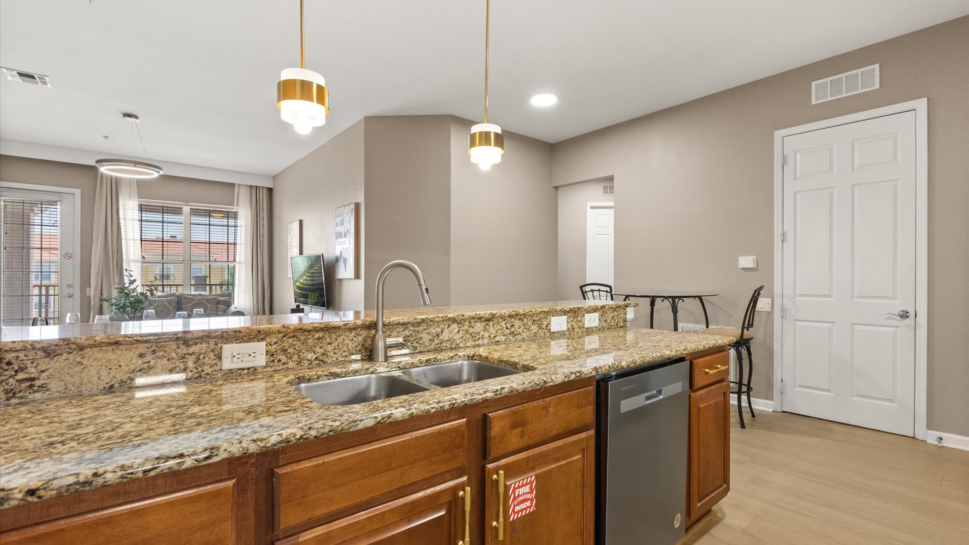 A view from the kitchen island looking towards the living and dining areas, highlighting the double sink set in granite countertops. Two modern pendant lights hang above the island, and light wood floors extend into the open-concept living space.