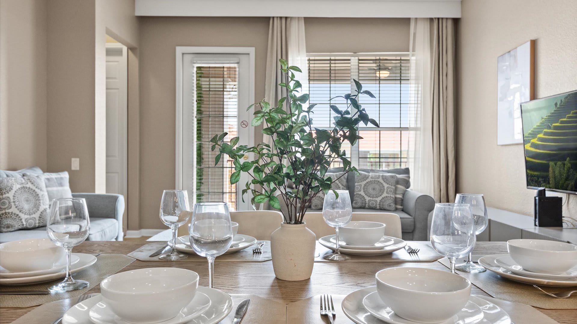 A close-up view of the dining table, meticulously set with white dinnerware, wine glasses, and a green plant centerpiece in a cream vase. The light wood table and beige upholstered chairs are visible, with a gray sofa and window blinds in the background.