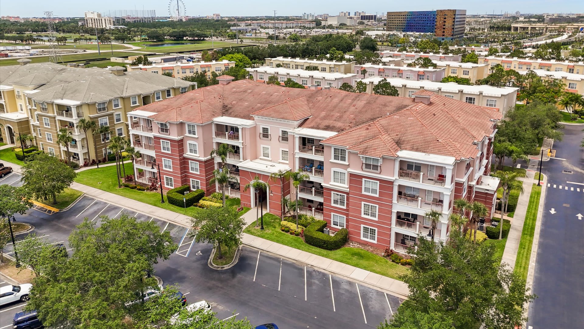Aerial view of a large, multi-story condominium building with a red tile roof and pink/red facade, surrounded by parking lots, green spaces, and other residential buildings under a cloudy sky.
