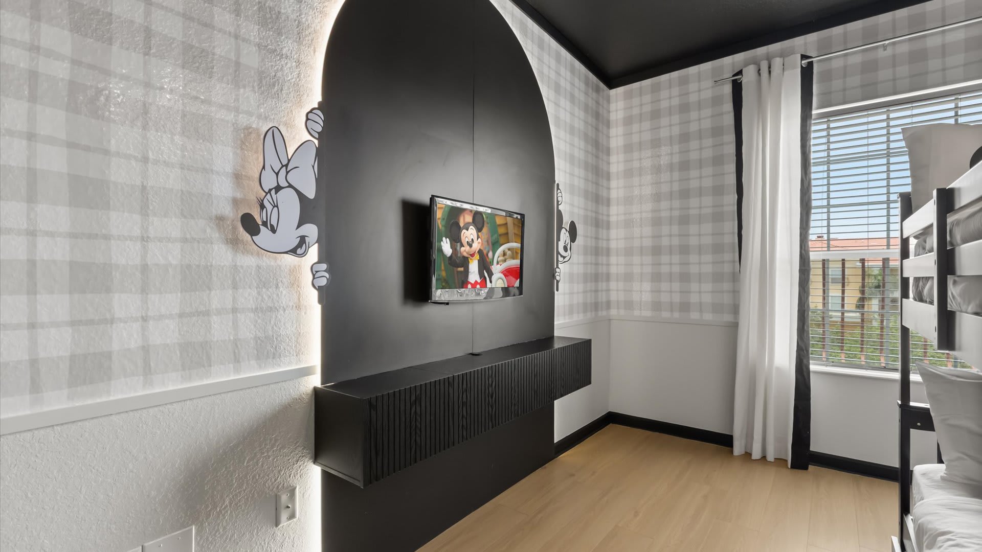 A close-up of the Mickey Mouse themed bedroom wall, featuring a wall-mounted television above a black floating console. A Minnie Mouse silhouette with backlighting adorns the plaid wallpaper.