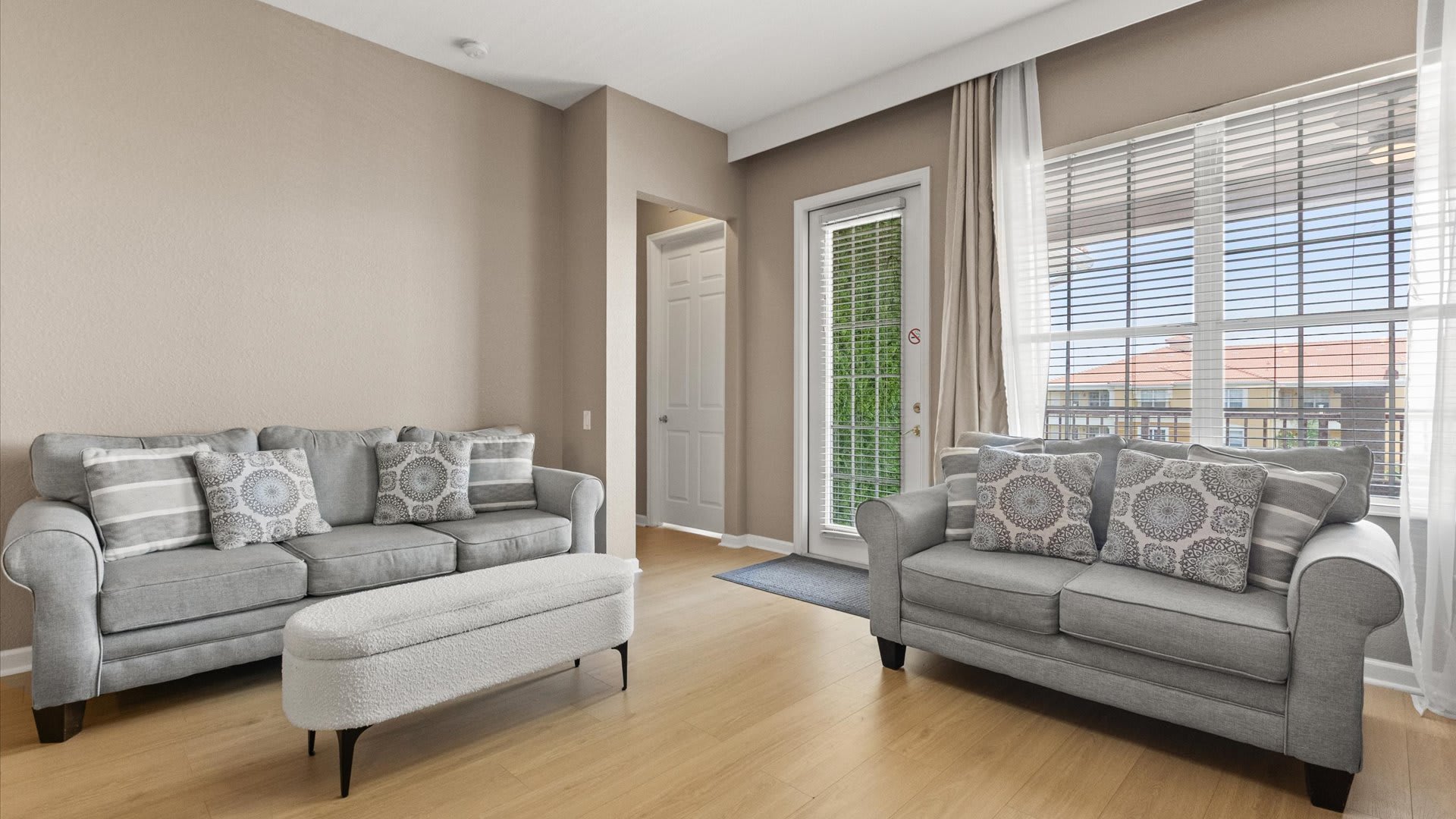 A cozy living room features two gray upholstered sofas adorned with patterned throw pillows, facing each other with a white textured ottoman in between. Light wood floors and neutral walls create a bright space, with a glass door leading to an outdoor area.