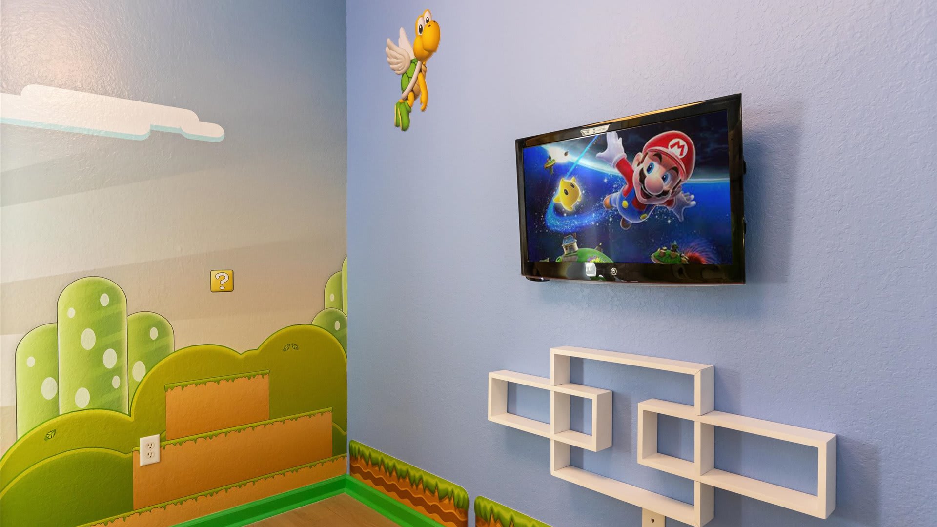 A section of the Super Mario themed bedroom showing a wall-mounted television above white floating geometric shelves. The blue wall features a flying Koopa Troopa figure and game-inspired graphics.