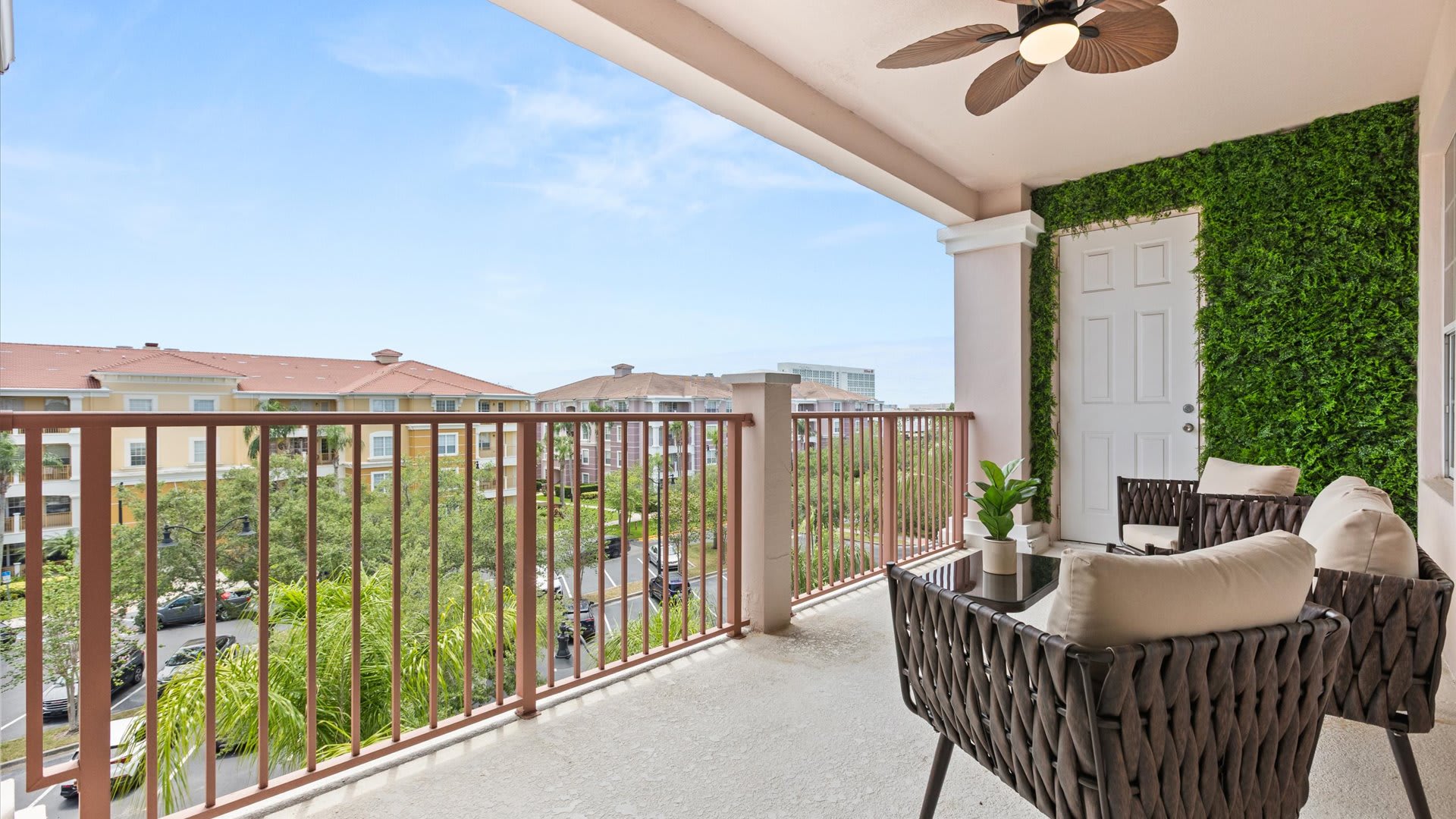 Spacious covered balcony with outdoor seating, a green artificial turf wall, and a view of the surrounding residential buildings and street below. A ceiling fan is installed overhead.