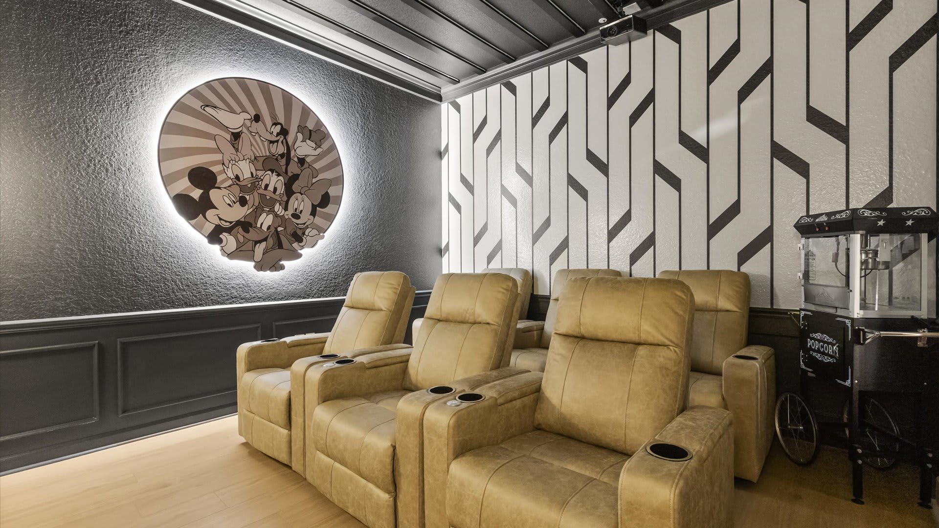 Wide view of the media room showcasing multiple tan leather recliners with cup holders, a vintage-style popcorn machine, and a backlit circular Disney character art piece on the textured wall.