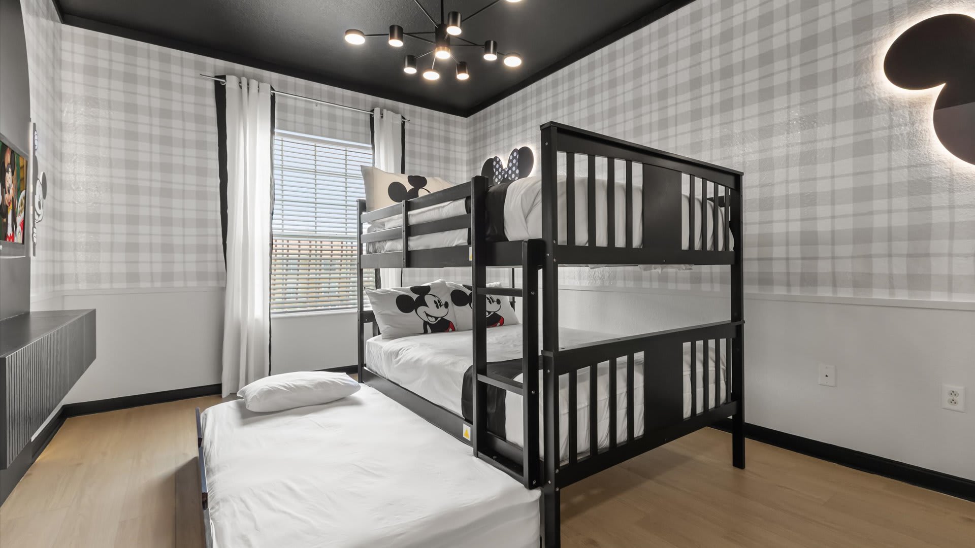 A full view of the Mickey Mouse themed bedroom, highlighting the black bunk bed with a trundle bed pulled out. The room features plaid wallpaper, a dark ceiling, and a window with white curtains.