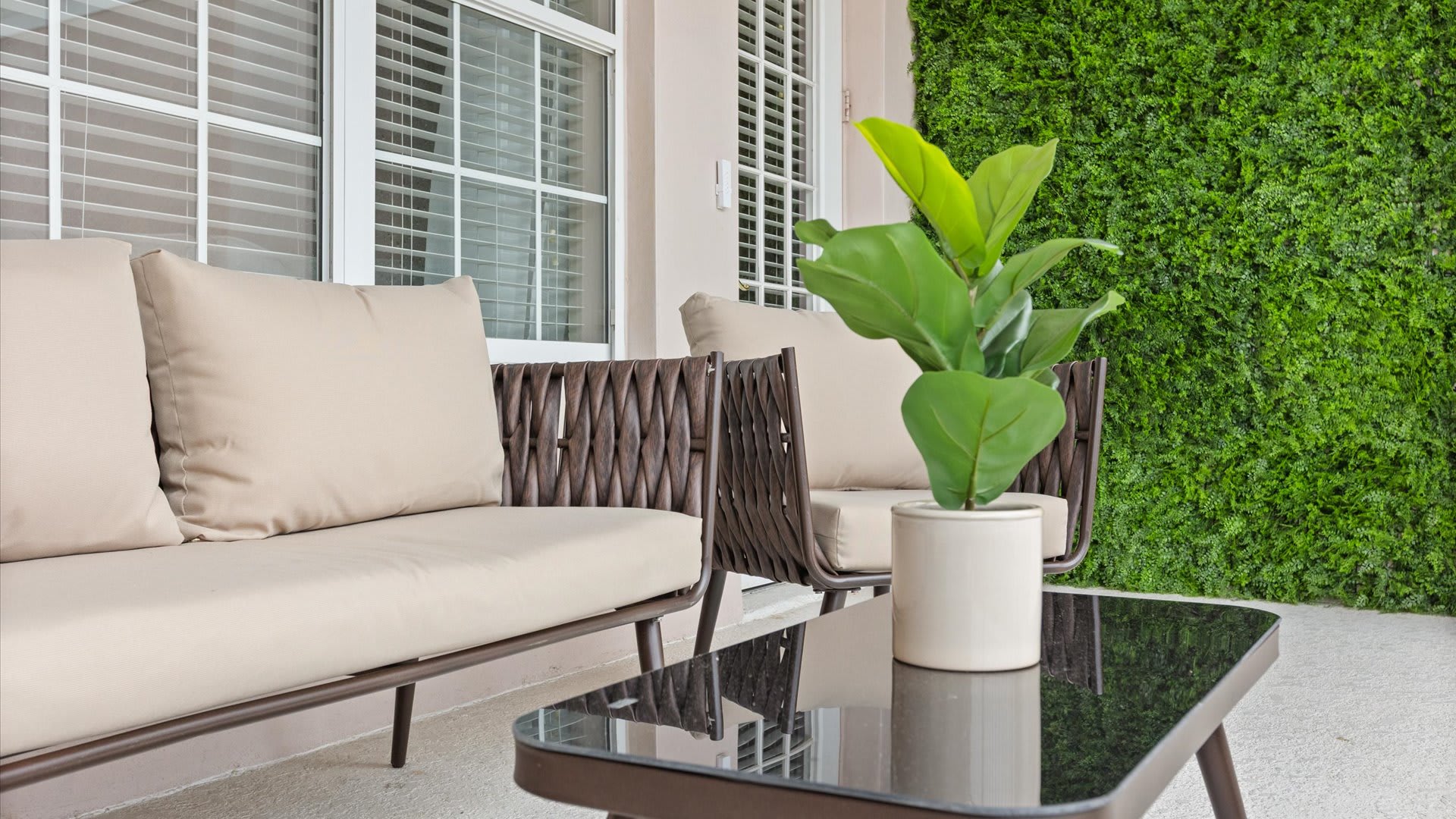 Close-up view of the outdoor seating on the balcony, featuring a brown woven sofa with beige cushions, a glass-top coffee table, and a potted plant.