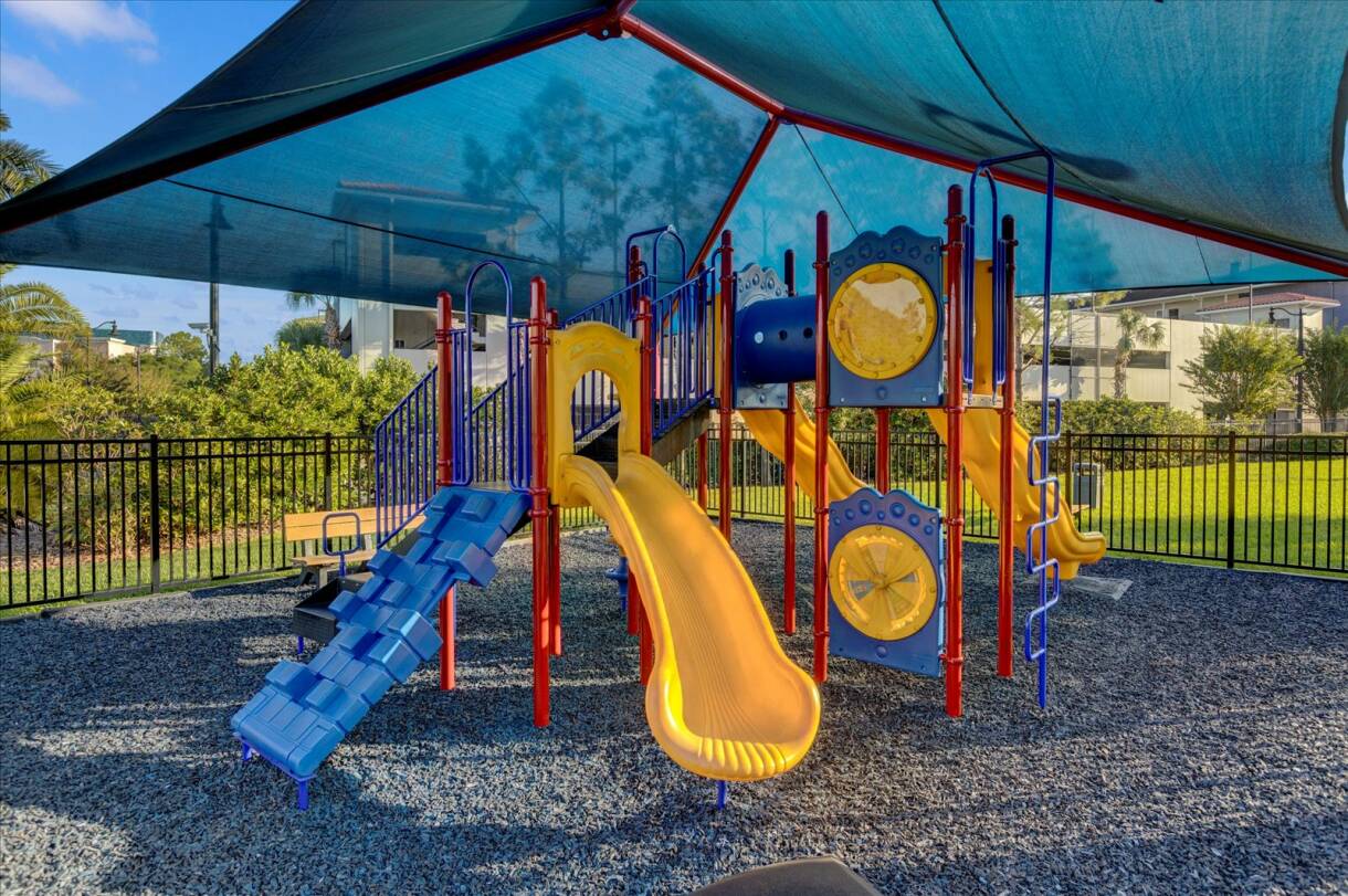 A vibrant outdoor children's playground features a multi-level structure with yellow slides, blue stairs, and climbing elements, all covered by a large blue shade sail. The ground is covered with soft pebbles.