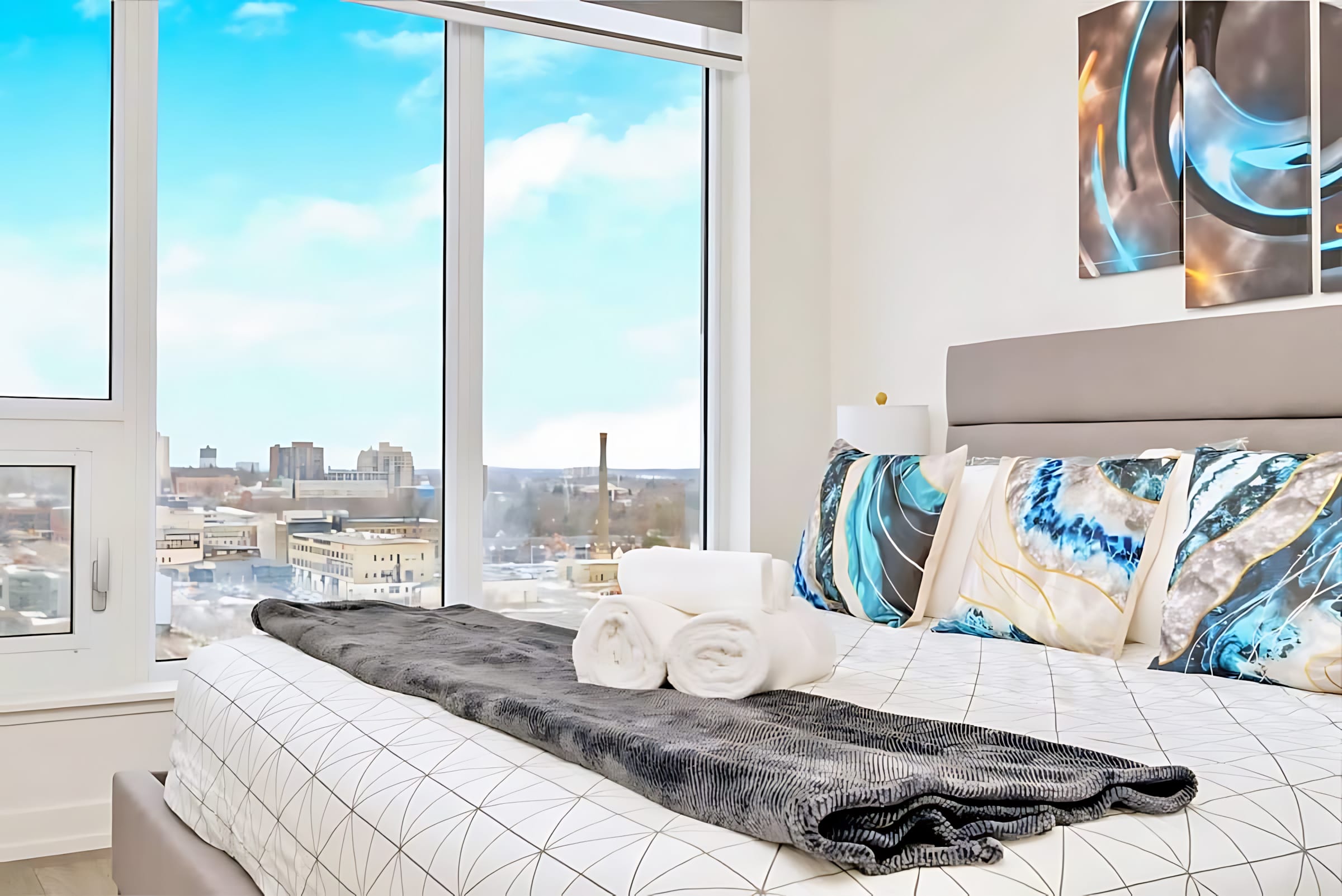 Bedroom with a queen-sized bed featuring geometric bedding and blue accent pillows, positioned next to a large window offering expansive city views. Rolled white towels are neatly placed on the bed.