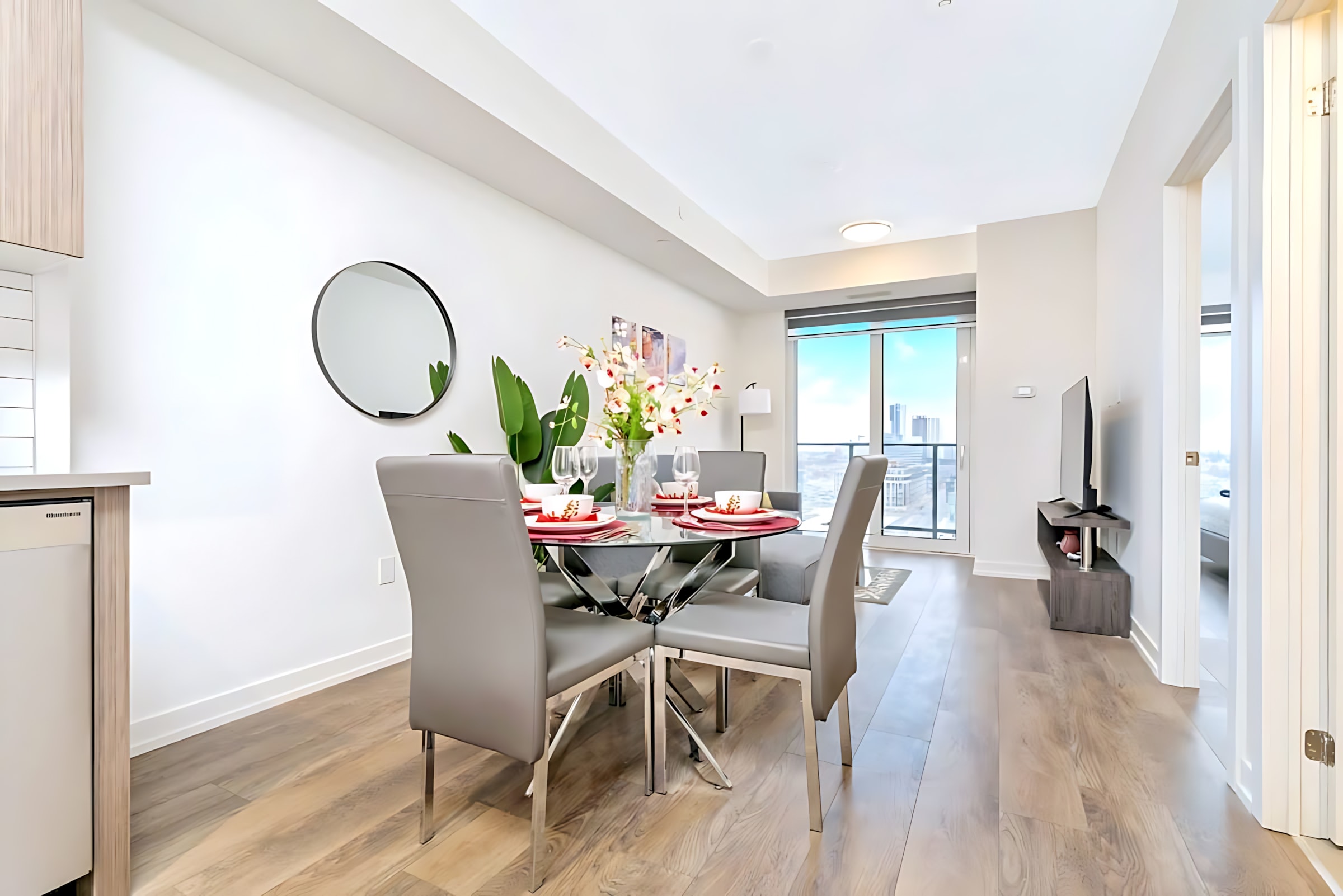 Bright open-concept dining area with a glass-top table and four grey upholstered chairs, leading into a living room with a large window offering city views and a wall-mounted TV.