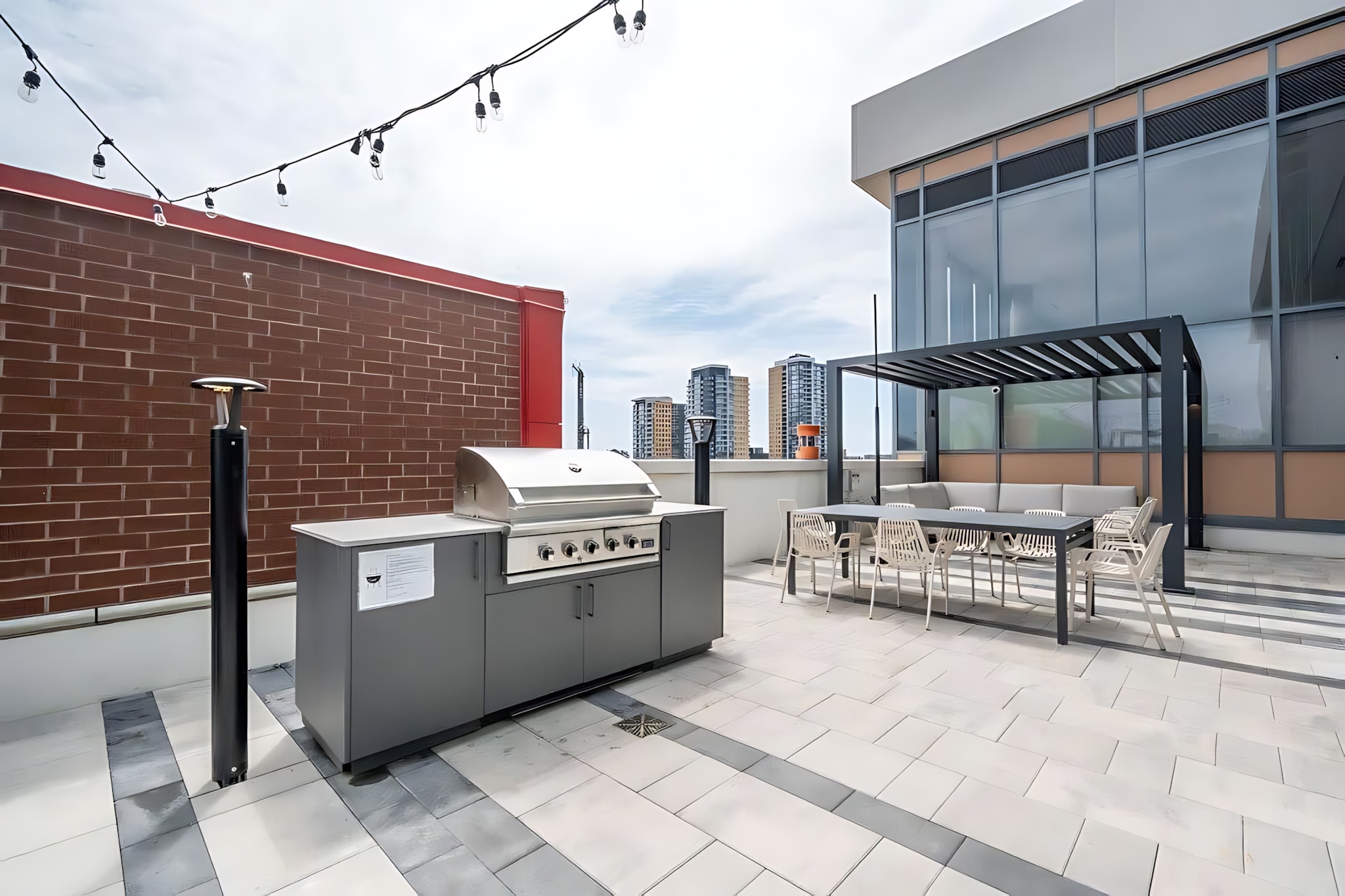 Expansive rooftop patio area featuring a large outdoor grill station, a dining table with chairs under a pergola, and string lights, all set against a backdrop of other city buildings.