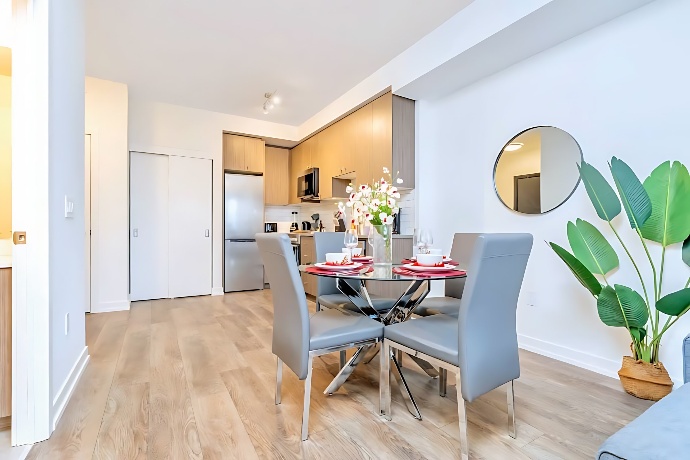 Modern open-concept dining area with a glass-top table and four grey upholstered chairs, adjacent to a compact kitchen featuring light wood cabinetry, stainless steel appliances, and a white subway tile backsplash.