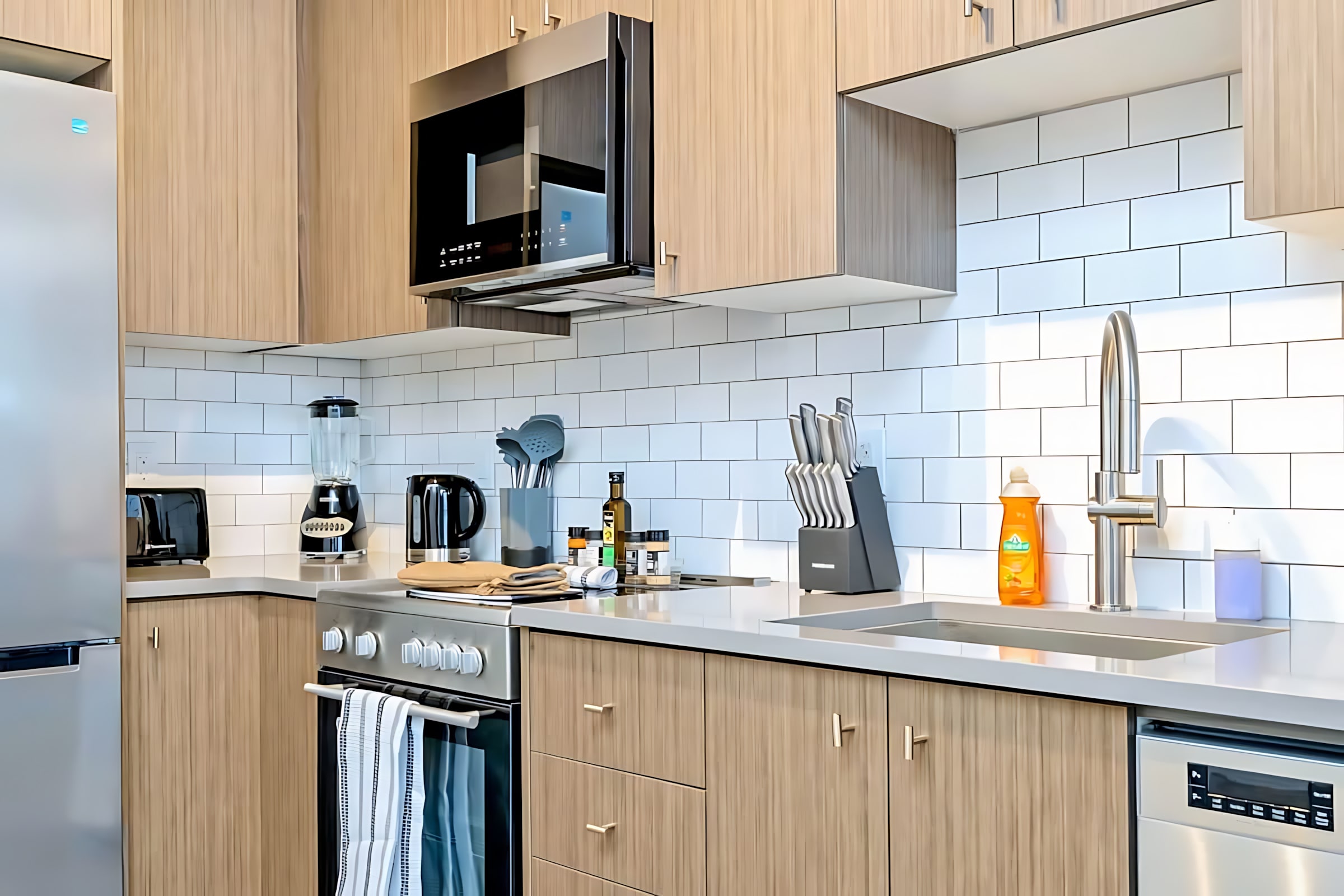 Detailed view of the modern kitchen counter with a stainless steel sink and faucet, white subway tile backsplash, various kitchen utensils, and small appliances like a blender and kettle.