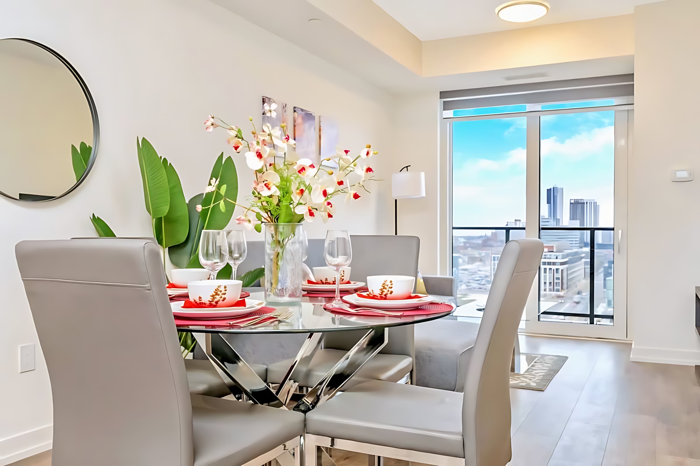 Elegant dining area with a round glass table set with white and red dinnerware, grey upholstered chairs, a floral centerpiece, and a large plant, with city views visible through the distant windows.