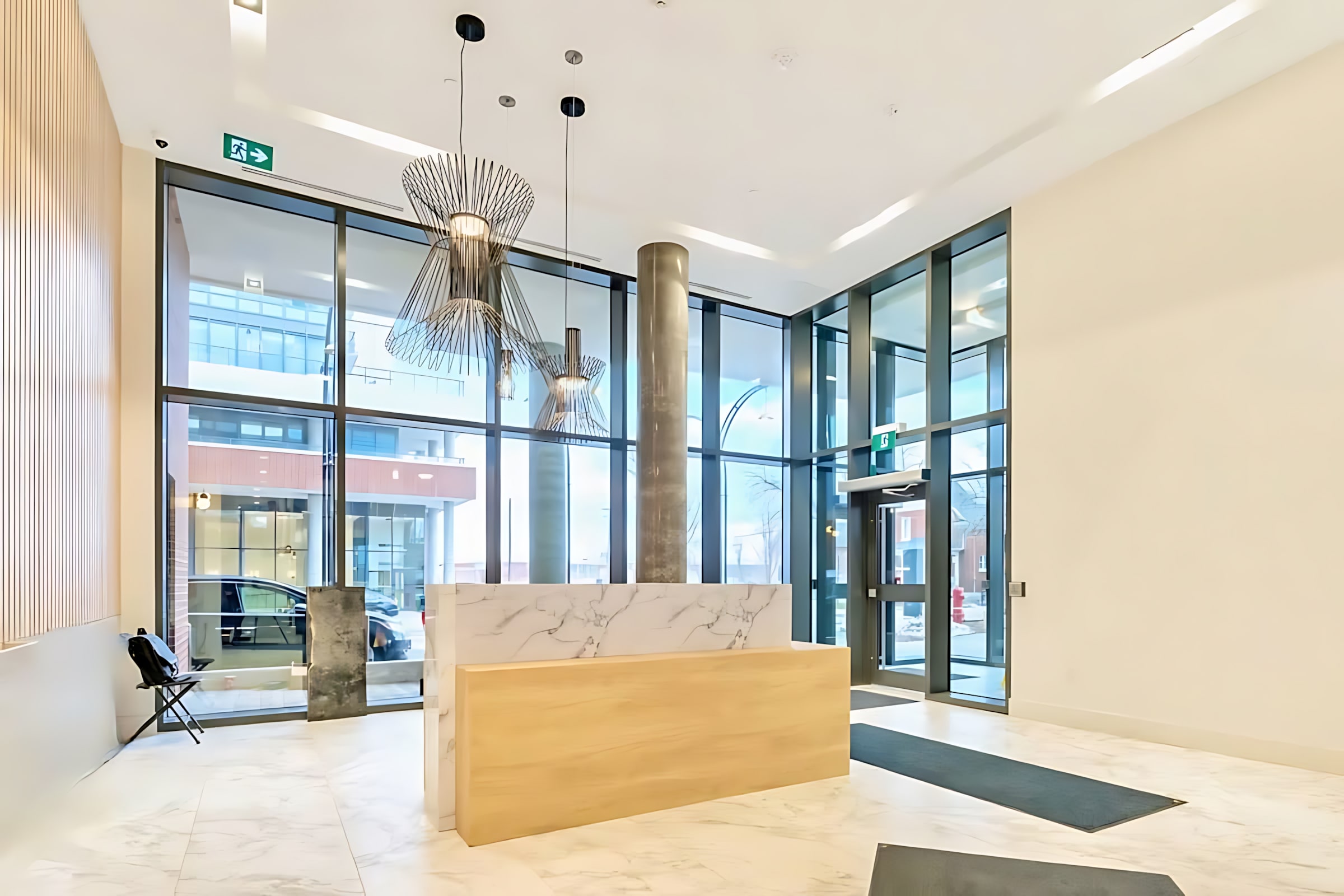 Bright and spacious building lobby with a modern reception desk featuring marble and wood finishes, large floor-to-ceiling windows, and unique wire-frame pendant lights.