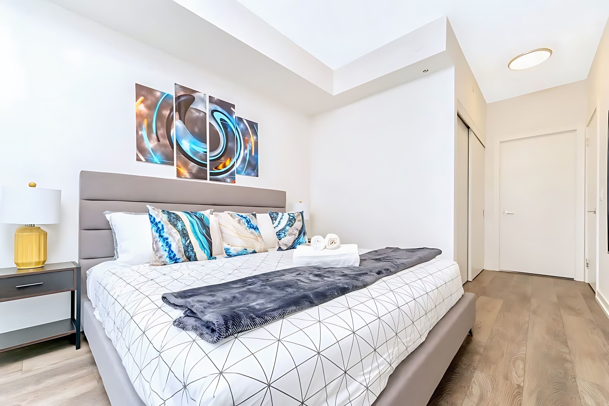 Contemporary bedroom featuring a queen-sized bed with a grey upholstered headboard, geometric pattern bedding, and blue and white decorative pillows, flanked by dark nightstands and a yellow lamp. Abstract multi-panel artwork adorns the wall above the bed.