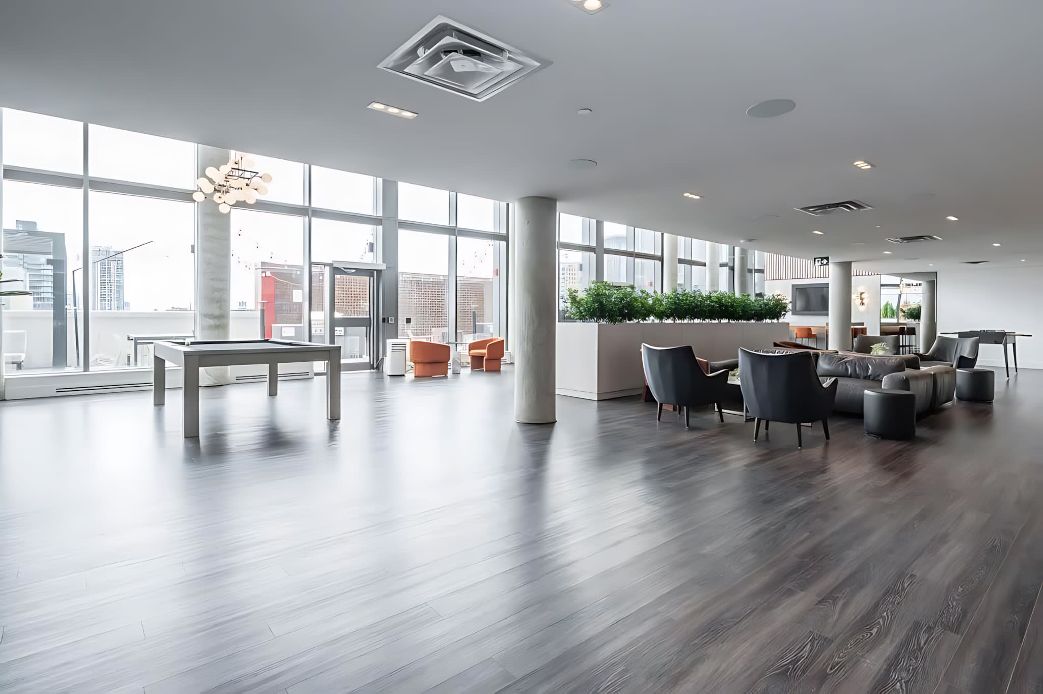 A spacious, bright common area features dark wood-look flooring and large floor-to-ceiling windows offering city views. A light-colored pool table occupies the foreground, while a lounge area with modern armchairs and a sofa is visible in the background, separated by a low planter with greenery.