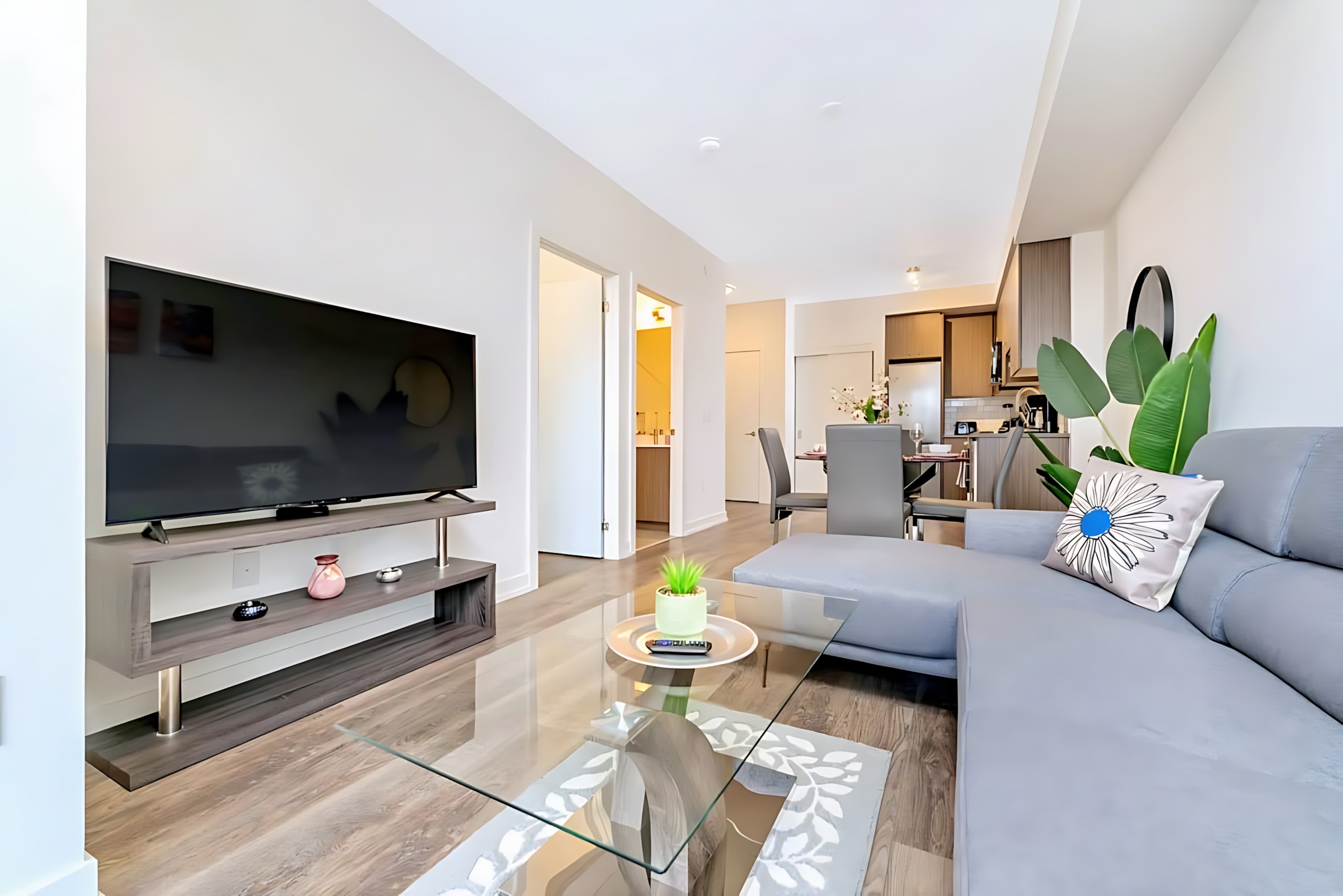 View of a modern living room with a large flat-screen TV on a grey media console, a grey sectional sofa with a decorative pillow, and a glass coffee table, with the kitchen and dining area visible in the background.