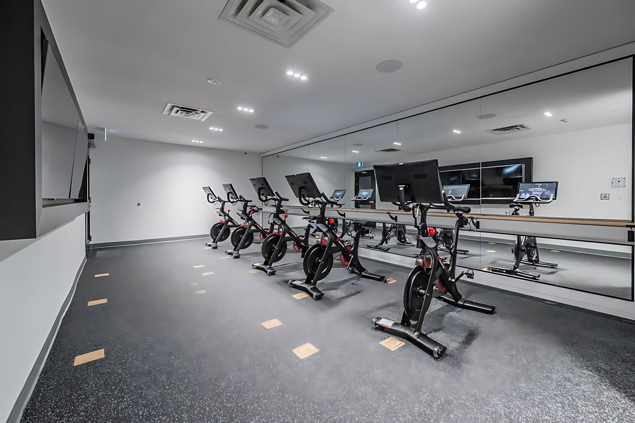 A modern fitness room features a row of six black exercise bikes with screens, facing a large mirrored wall that reflects the room. The space has a dark speckled floor, white walls, and recessed lighting, creating a sleek and functional workout area.