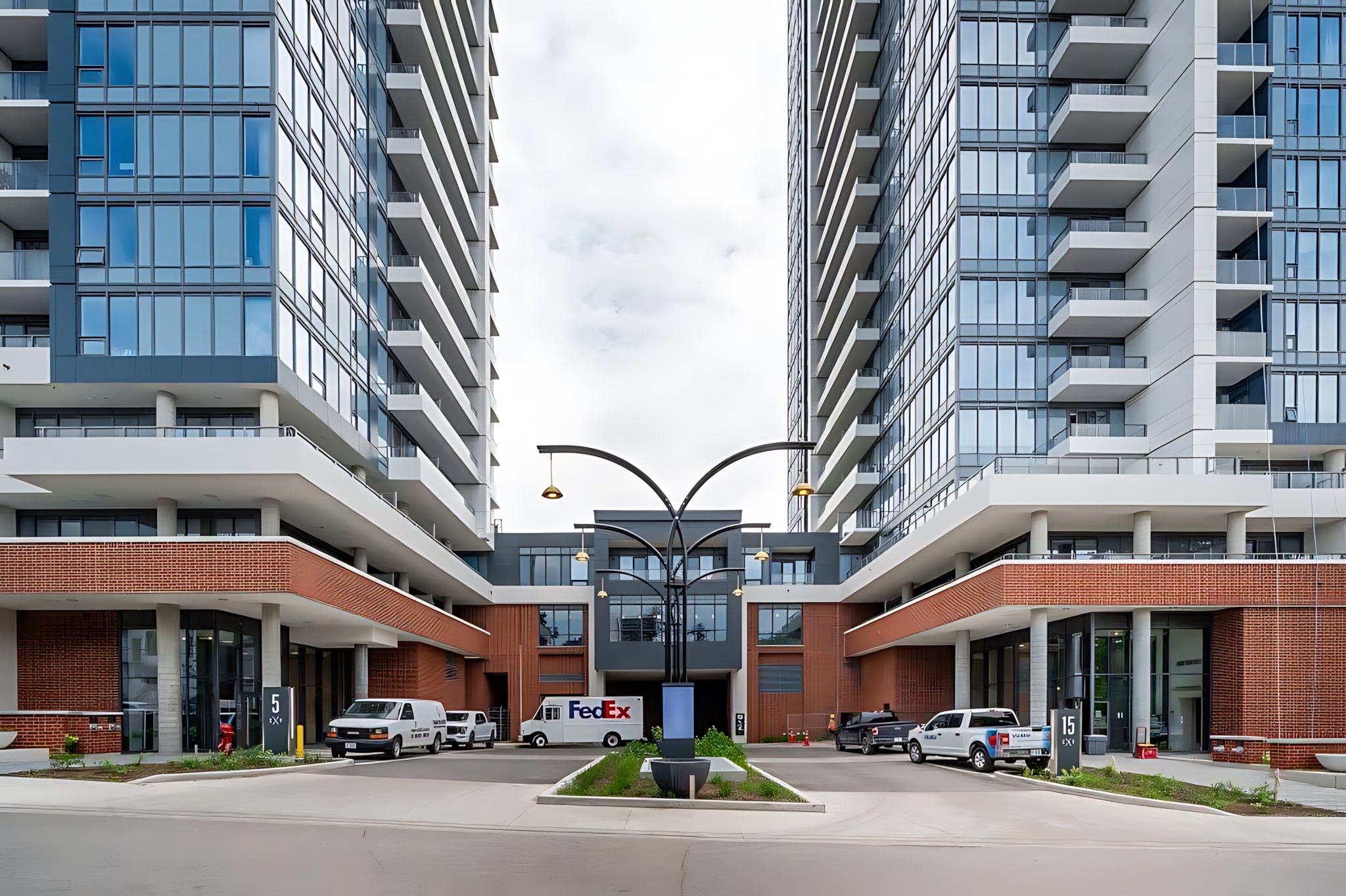 Modern high-rise apartment building exterior with multiple floors of glass windows and balconies, featuring a central drive-through entrance with brick accents and several parked vehicles.