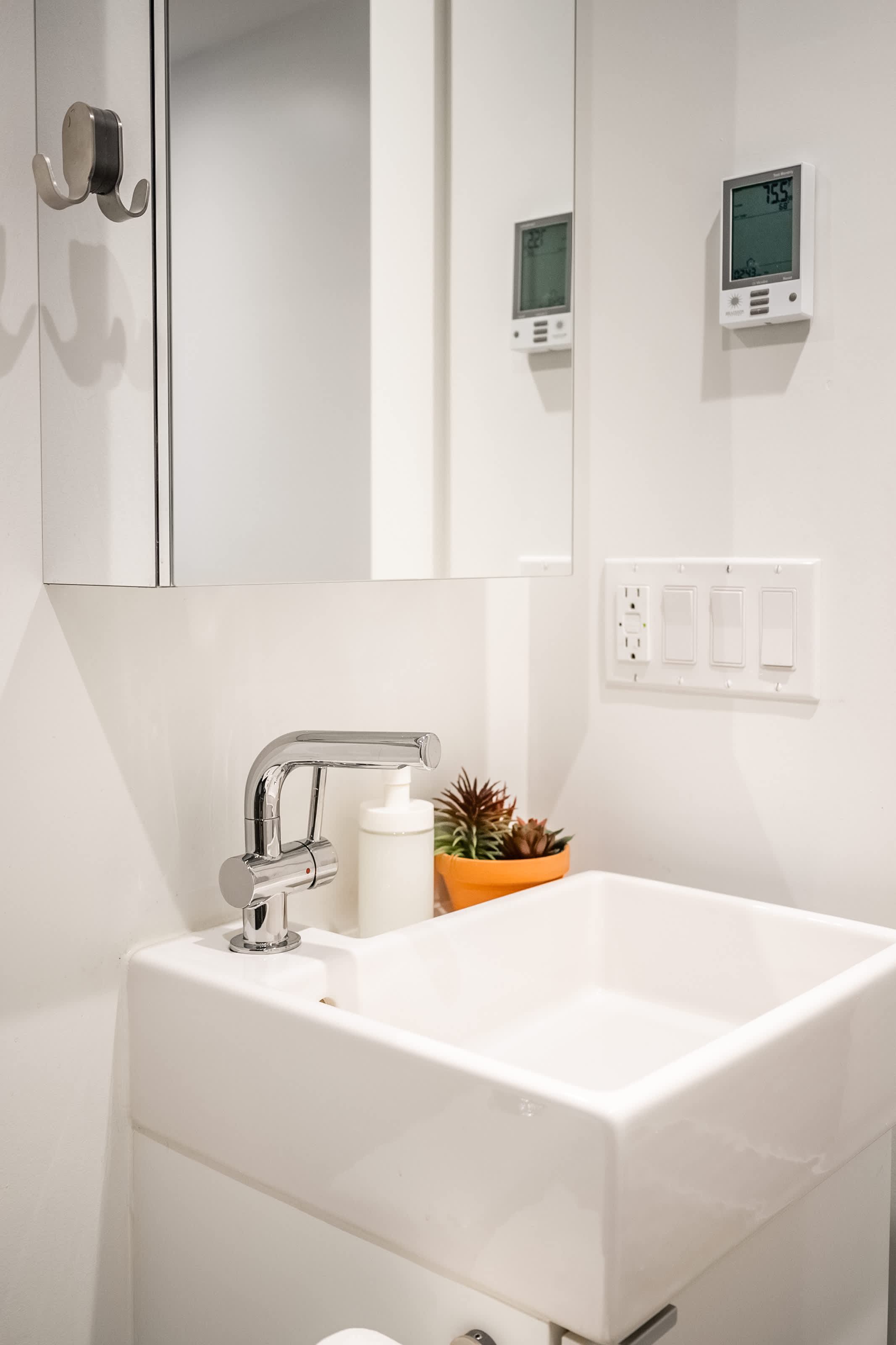A modern bathroom vanity with a square white sink, chrome faucet, and a wall-mounted medicine cabinet, featuring a small potted succulent and a soap dispenser on the counter, with light switches and a thermostat visible on the wall.