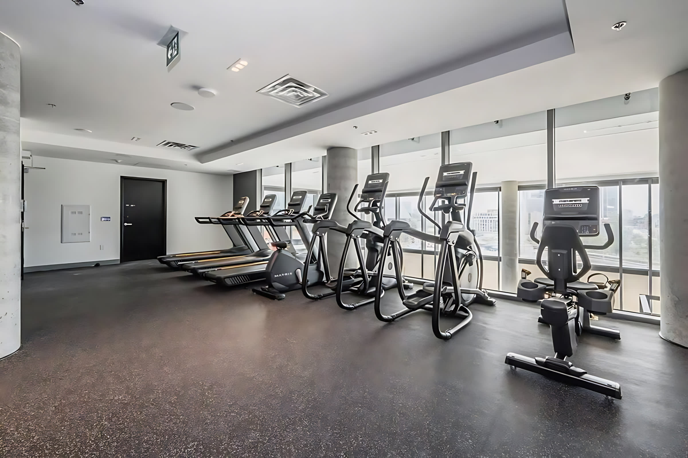Bright fitness center with a row of treadmills, elliptical machines, and stationary bikes on a dark rubber floor. Large windows provide natural light and city views, while white walls and pillars maintain a clean, modern aesthetic.