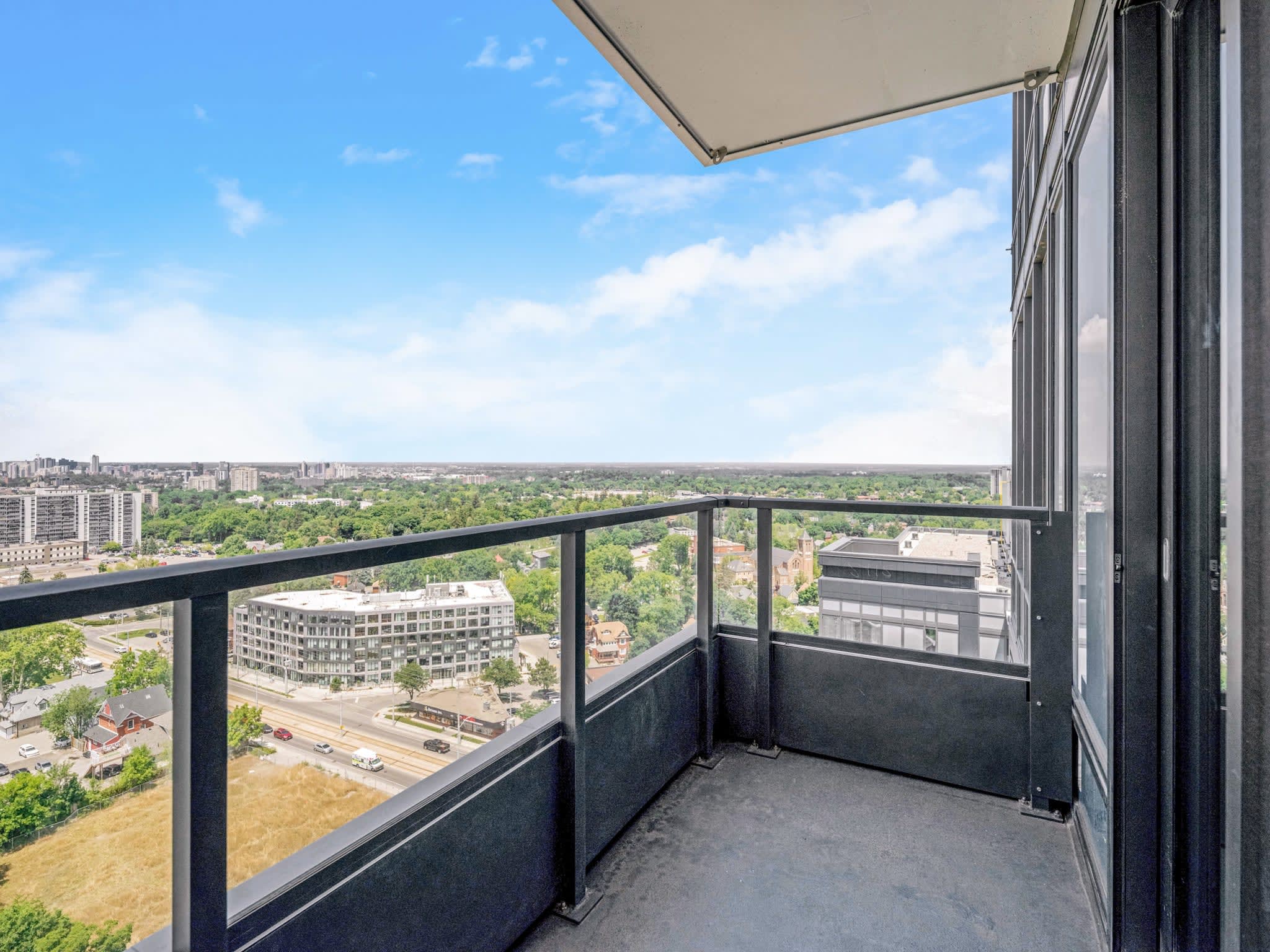 A spacious balcony with a black metal and glass railing provides an expansive panoramic view of a city, featuring a mix of high-rise buildings, residential areas, and lush green trees under a bright blue sky. Part of the building's exterior is visible on the right.