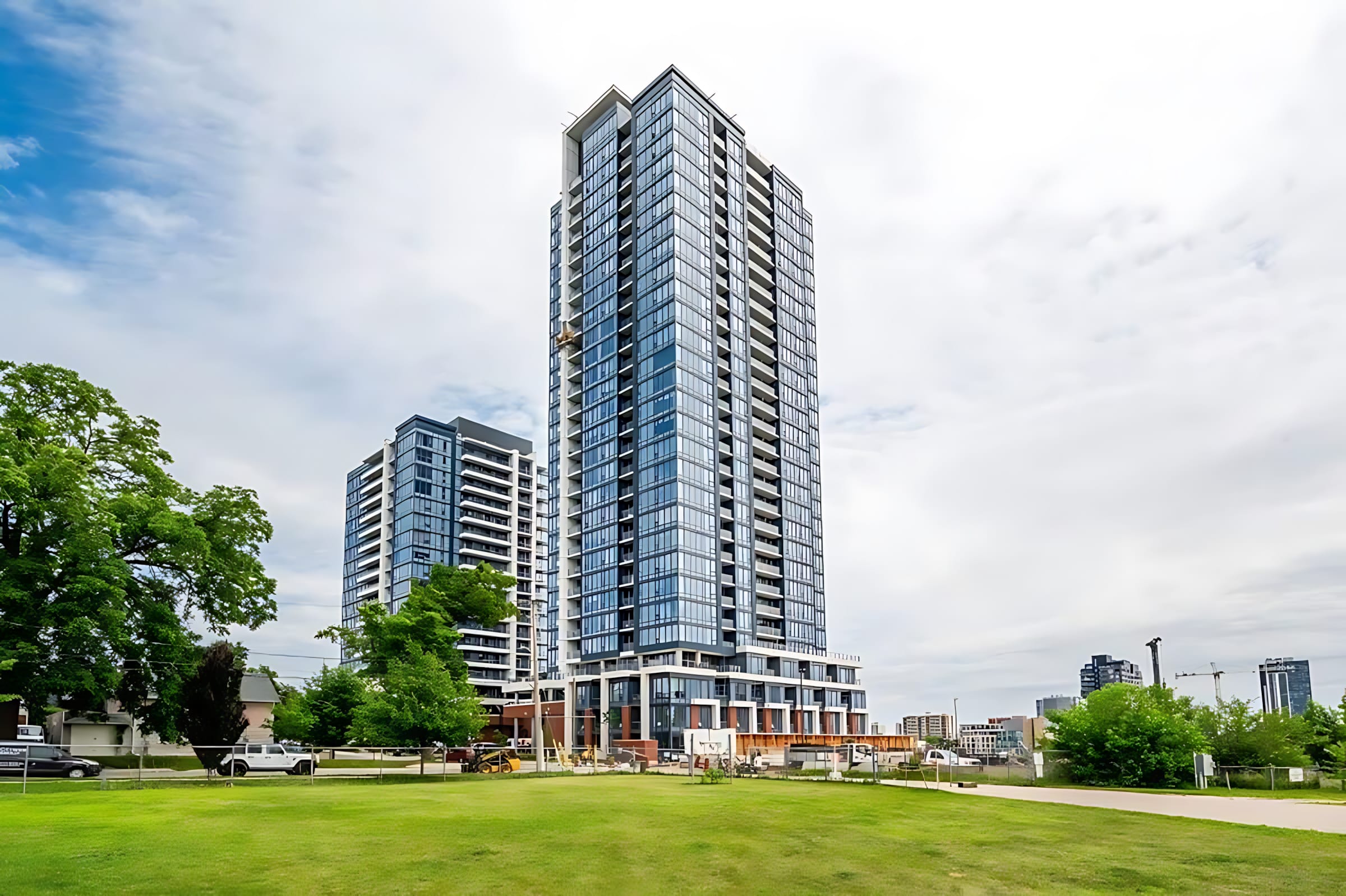 A tall, modern high-rise building with a glass facade stands prominently against a cloudy sky, surrounded by green grass and trees, with another building visible in the background.