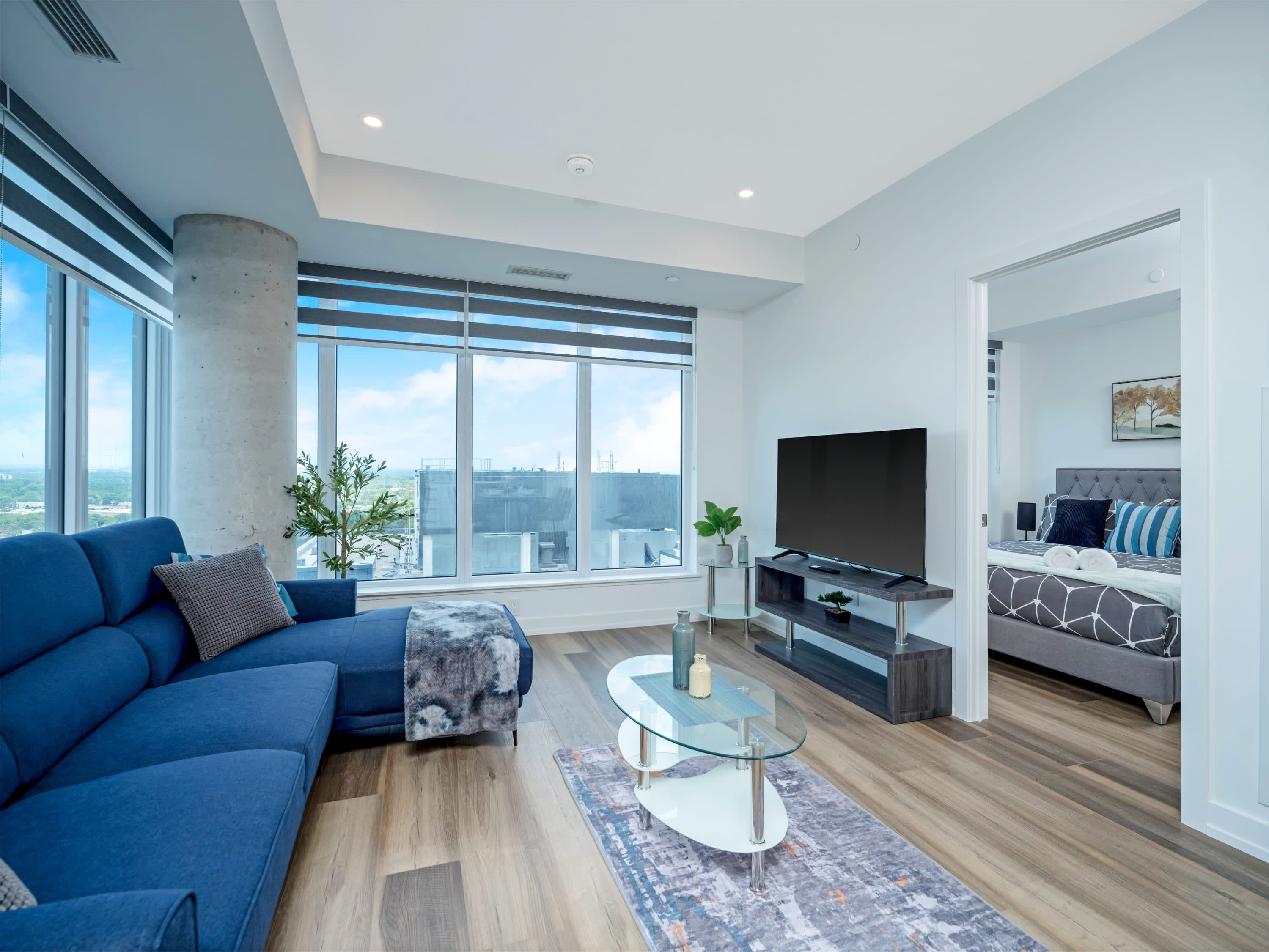 The modern living room features a blue sectional sofa, a glass coffee table on a patterned rug, and a wall-mounted TV on a dark media console. Floor-to-ceiling windows with blinds provide ample natural light and a glimpse of the city outside.