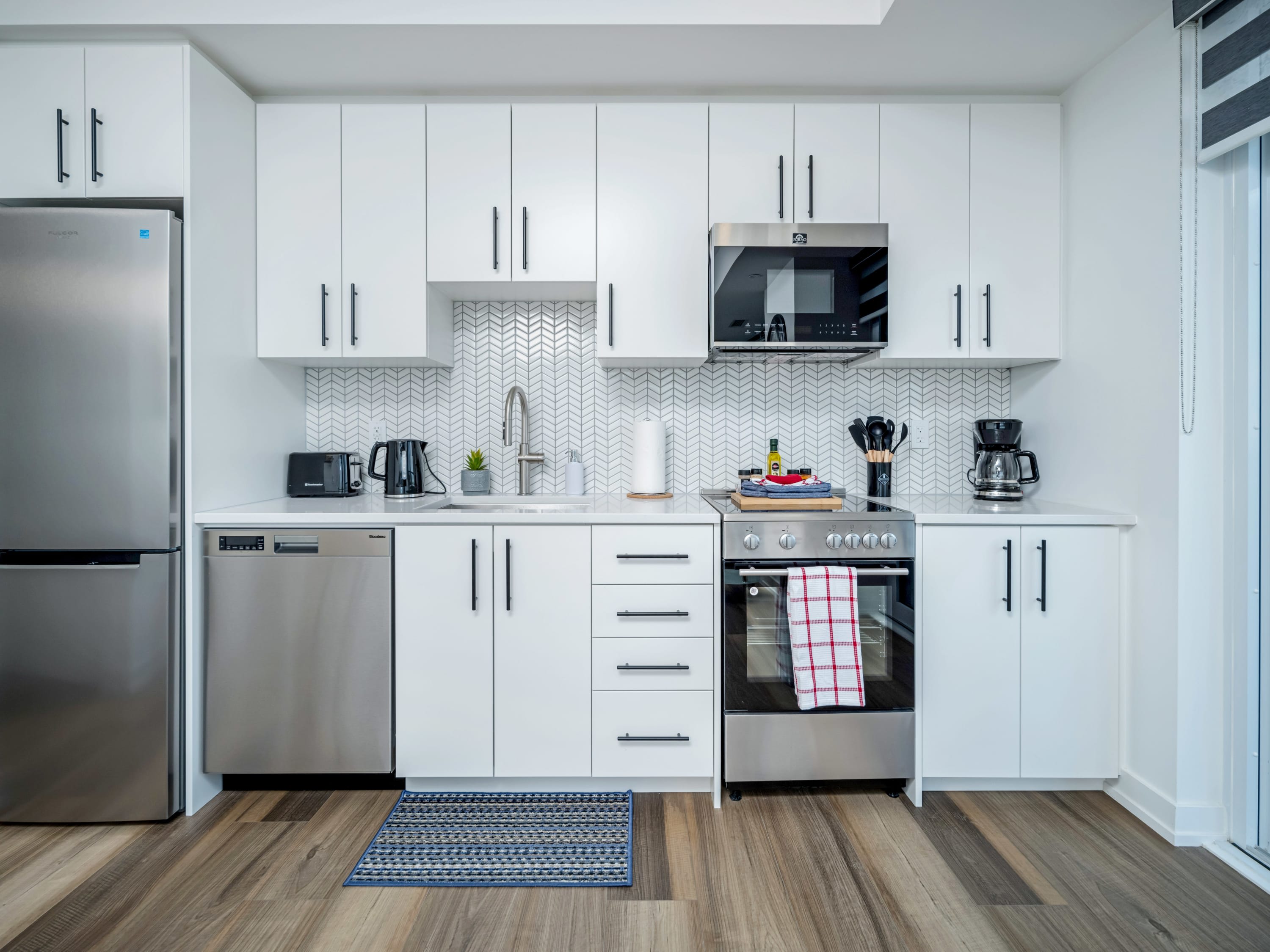 A bright, modern kitchen features white shaker-style cabinets, stainless steel appliances including a refrigerator, dishwasher, oven, and microwave, and a white herringbone tile backsplash.