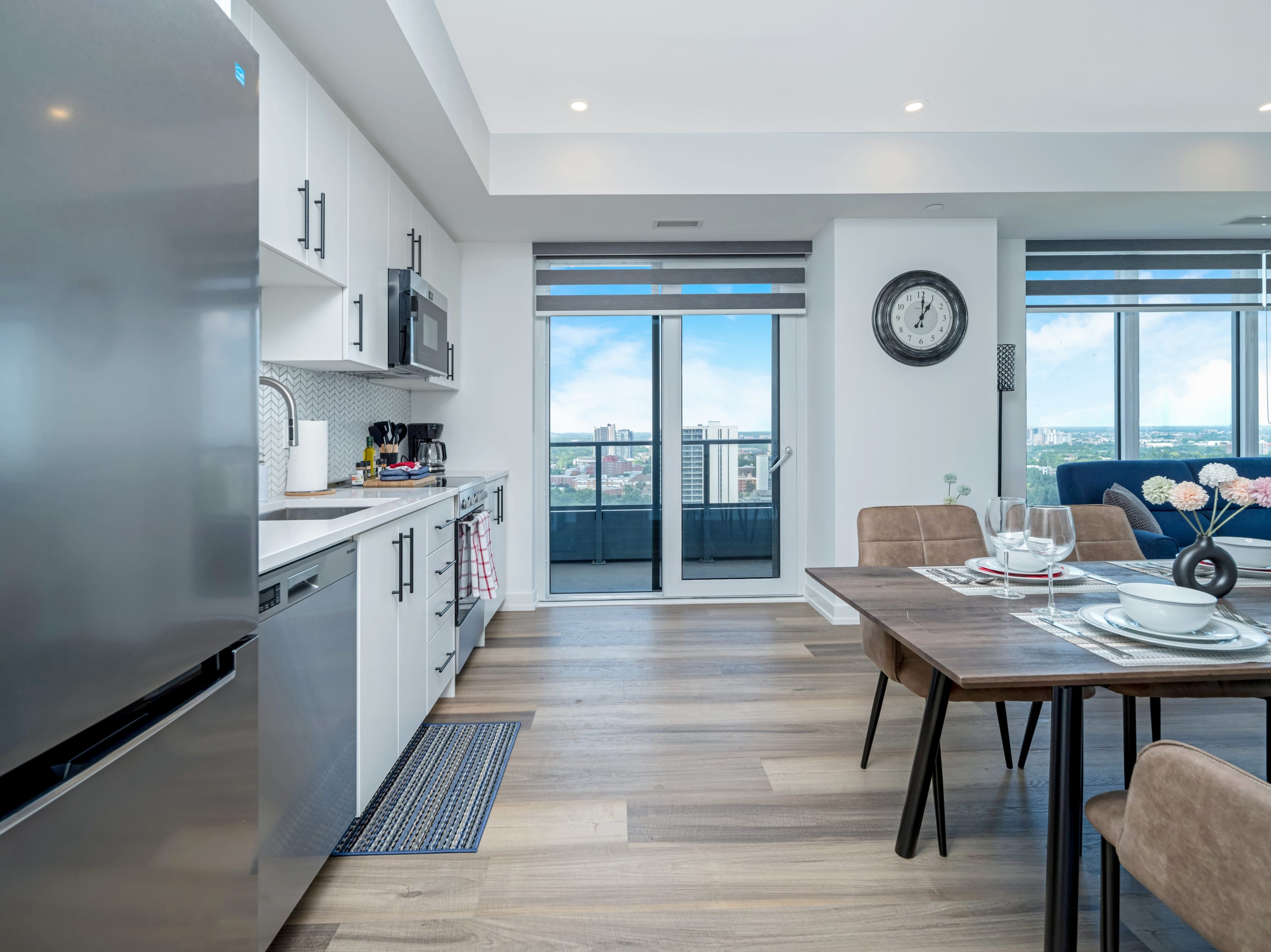 A modern kitchen with white shaker-style cabinets, stainless steel appliances including a refrigerator, dishwasher, and microwave, and a white subway tile backsplash. A wooden dining table with brown chairs is adjacent, and a glass door leads to a balcony.