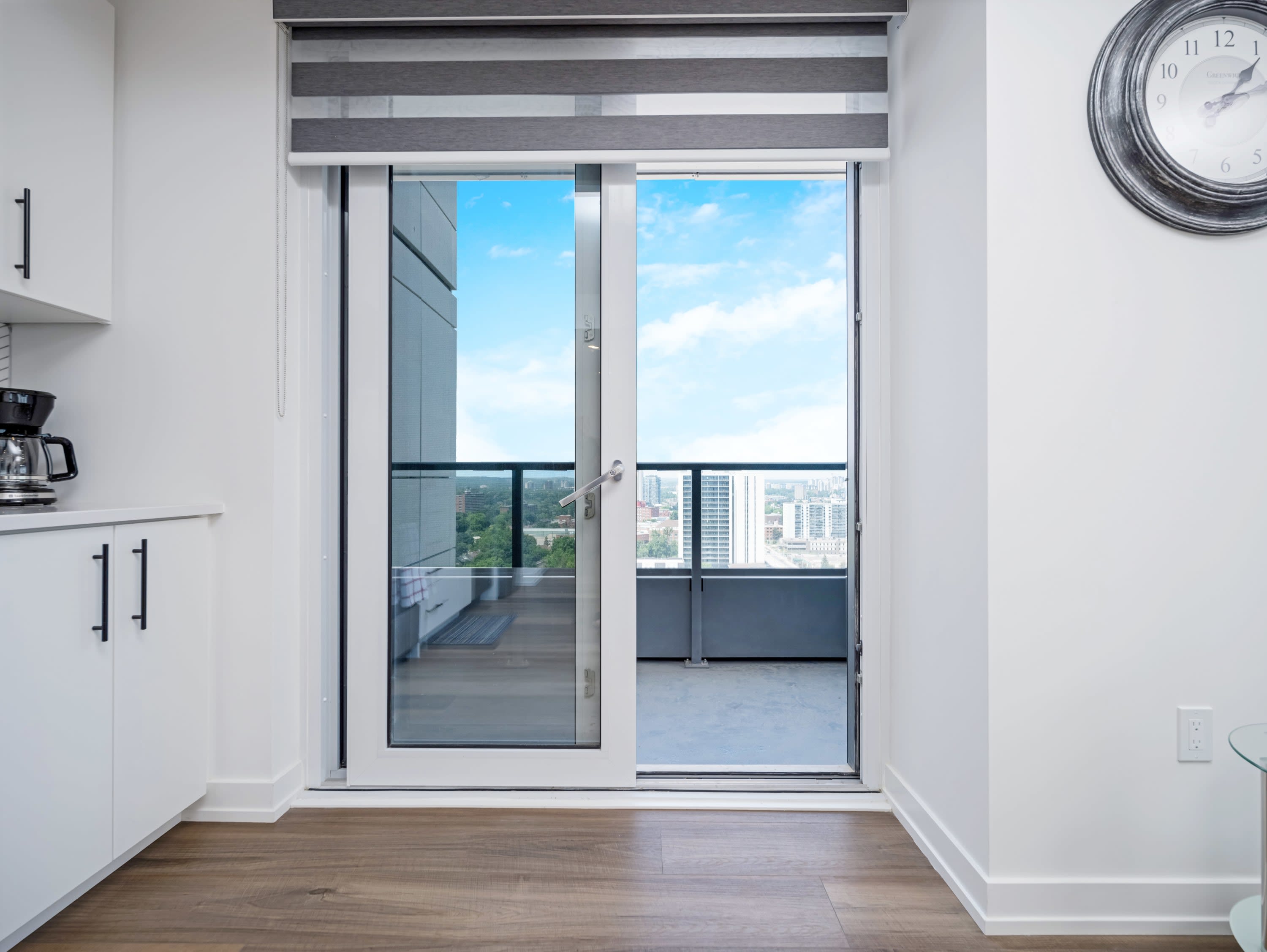 This image shows a glass door leading to a balcony with a black metal and glass railing, offering a clear view of the city below. The balcony floor is dark, and the sky is bright blue with scattered clouds.