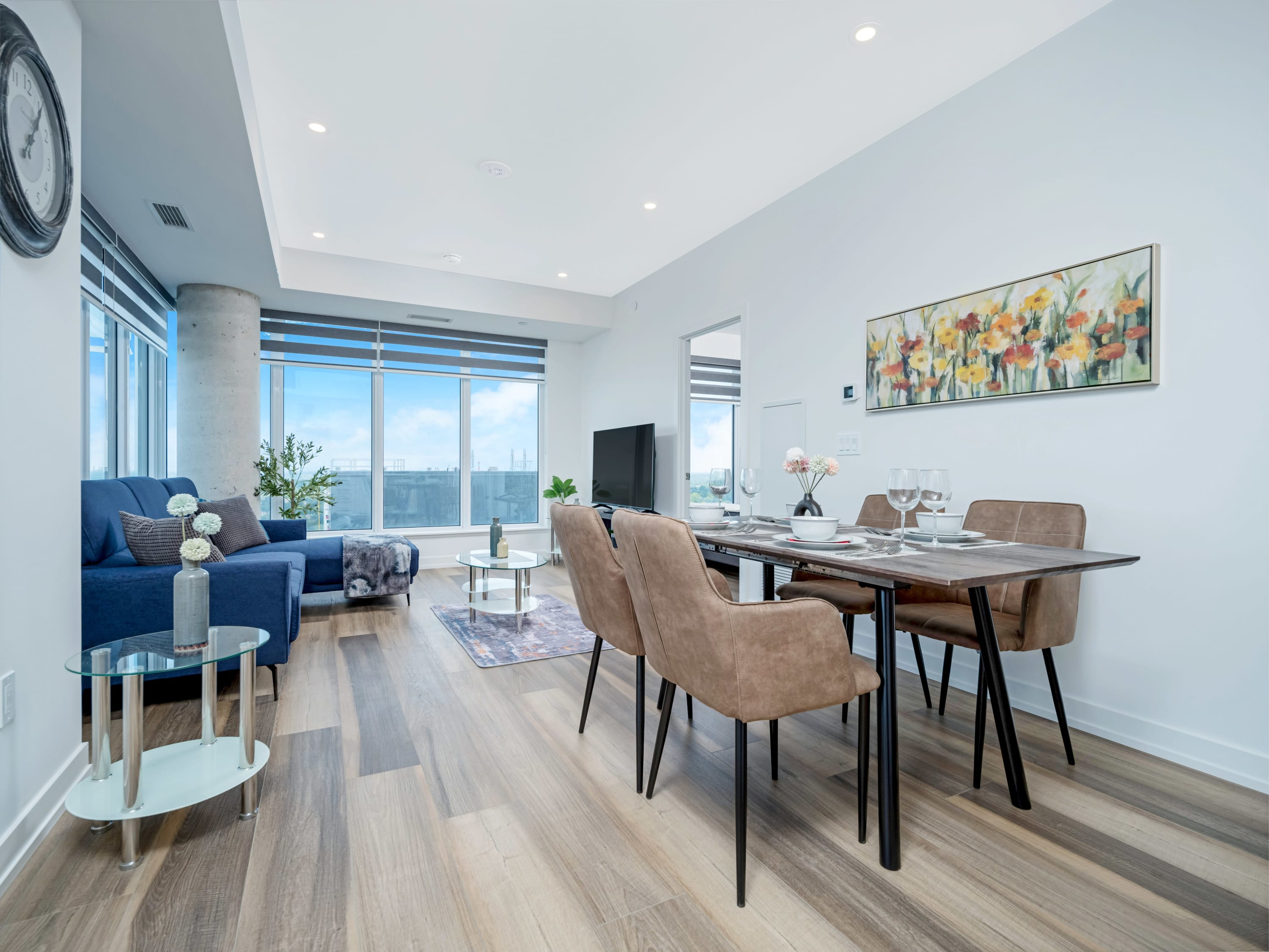 This open-concept living and dining area features light wood-look flooring, a blue sectional sofa, and a glass coffee table on a patterned rug. A wooden dining table with four brown chairs is set for a meal, and large windows provide natural light and city views.