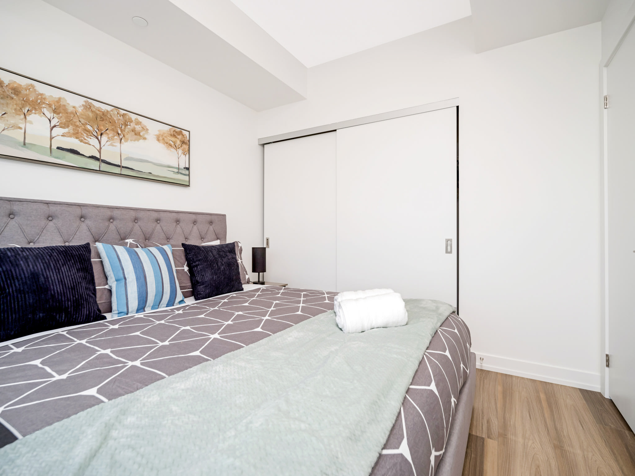 A cozy bedroom features a large bed with a grey upholstered headboard and geometric patterned bedding, flanked by a white sliding closet door and a small nightstand with a lamp.