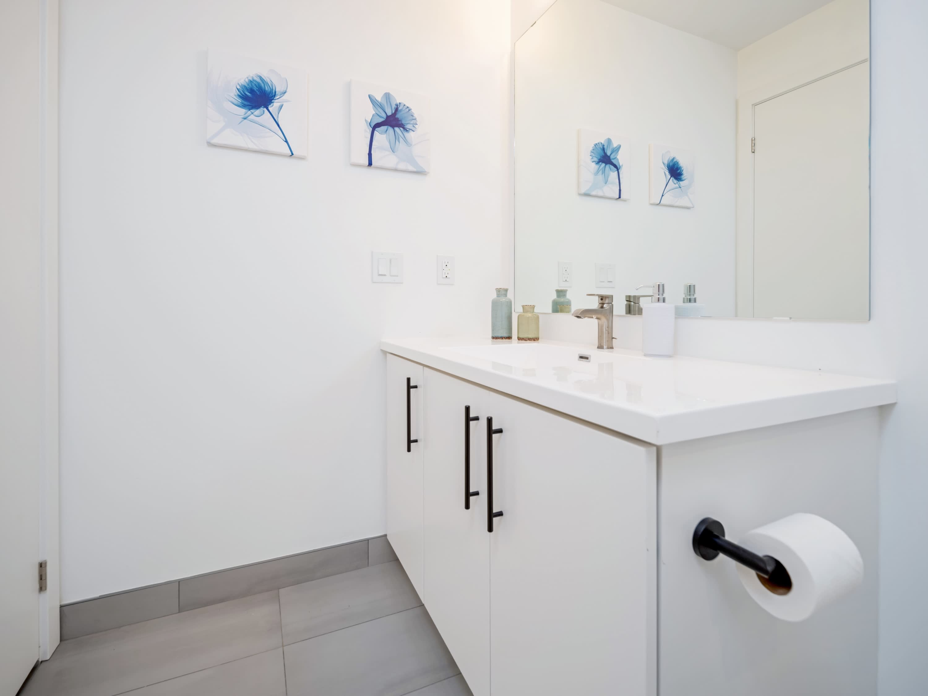 This modern bathroom features a white vanity with a large mirror, a sleek faucet, and three blue floral art pieces on the white wall, with grey floor tiles.