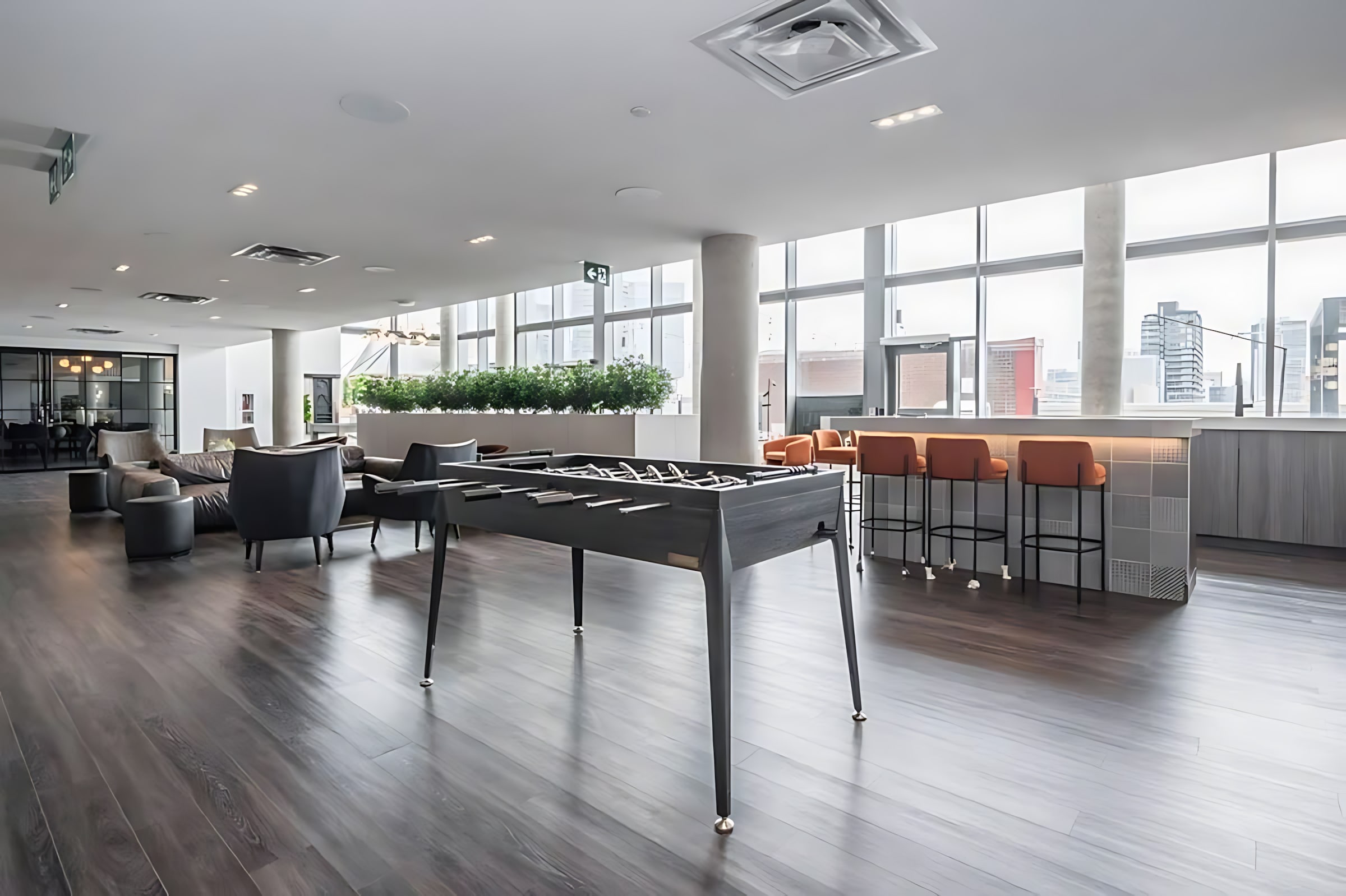 Modern common area featuring a dark foosball table on dark wood floors, surrounded by grey lounge seating and a bar with orange stools. Large windows offer city views, and a long planter with greenery adds a touch of nature.