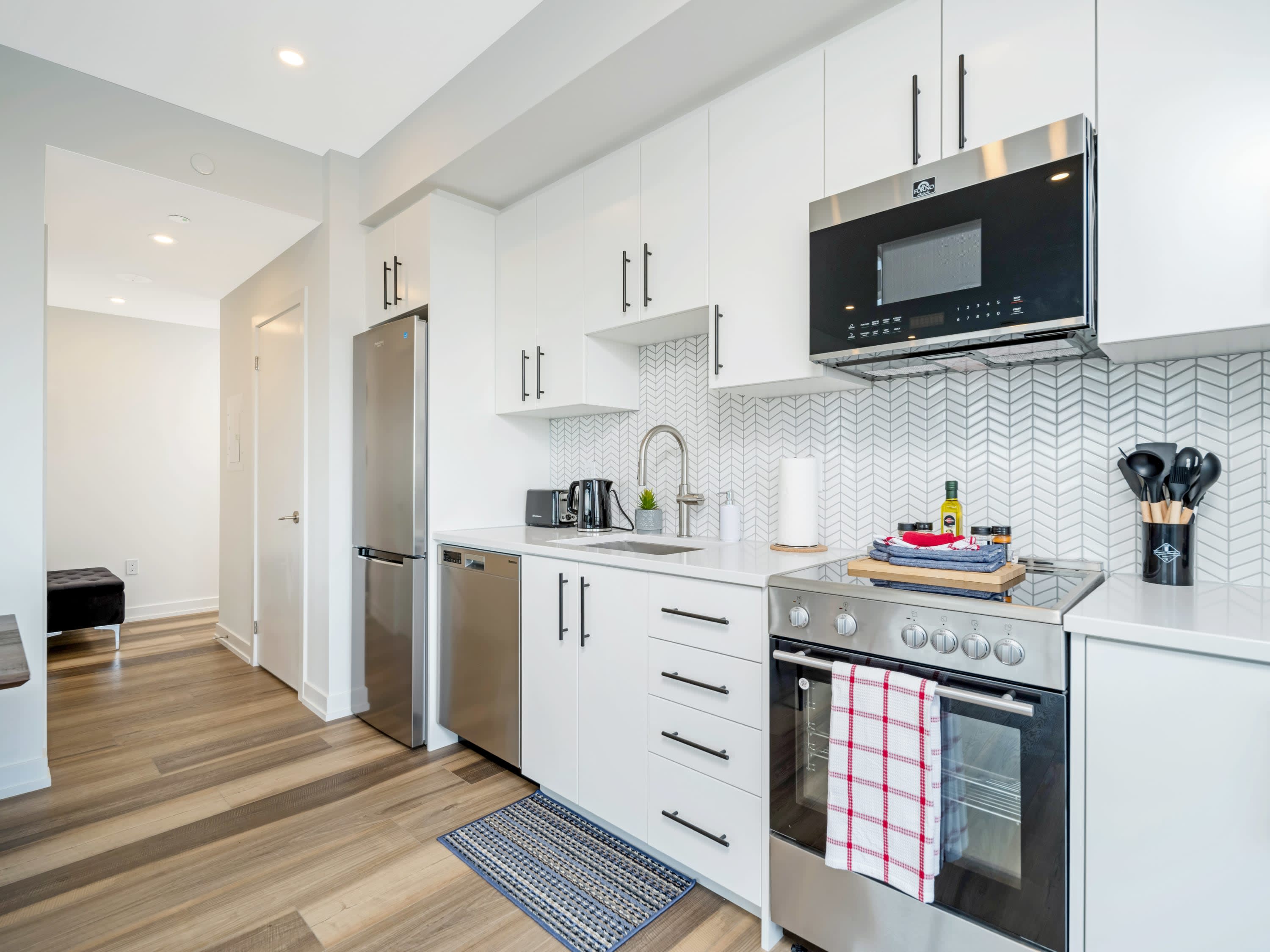 This view of the modern kitchen highlights the stainless steel appliances, white cabinetry with black hardware, and a herringbone tile backsplash, with light wood flooring extending into the hallway.